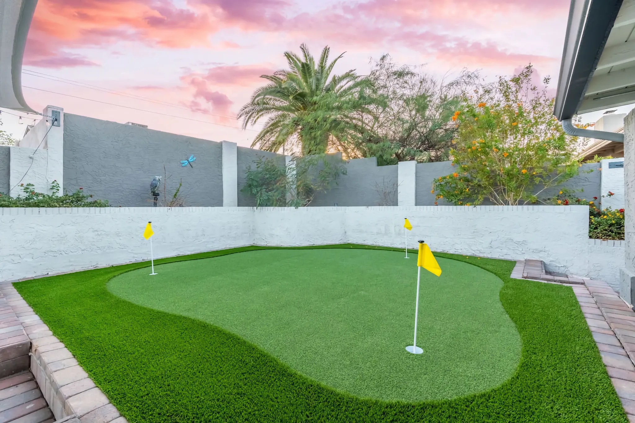 A lush artificial turf putting green with three yellow flags is situated in a walled backyard under a dramatic pink and orange sunset.