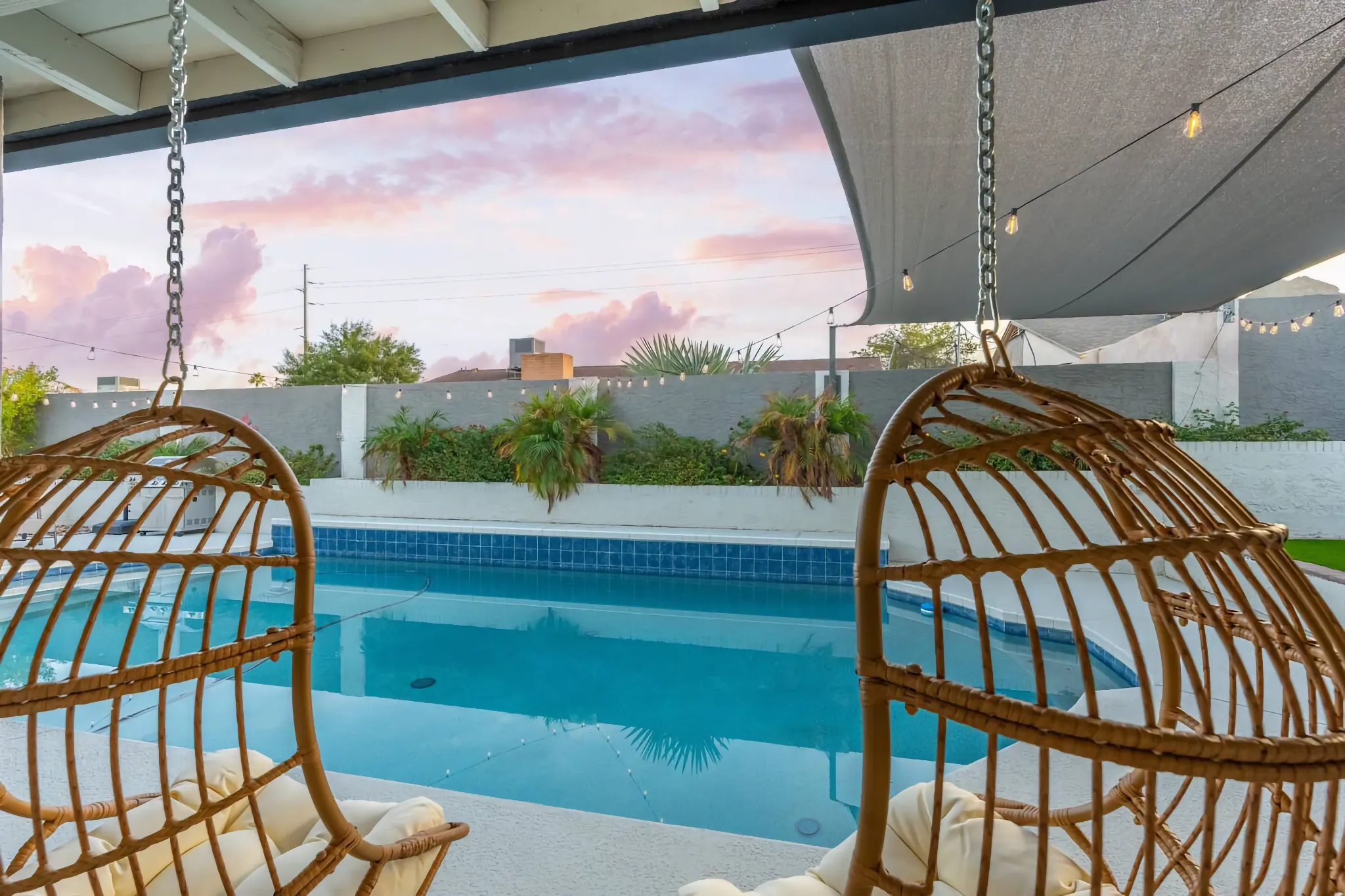 Two wicker hanging chairs frame a pool deck featuring a blue pool and string lights. Pink clouds illuminate the sky, creating a relaxing twilight atmosphere.