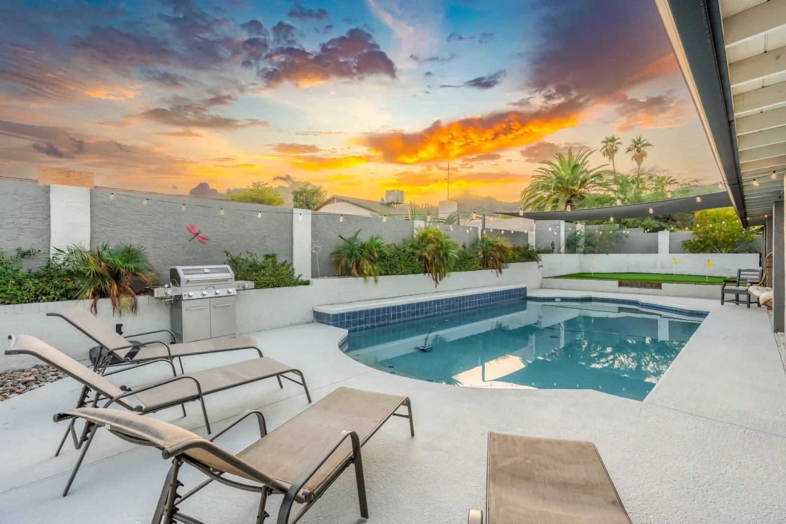 A backyard pool and patio with lounge chairs under a dramatic sunset sky of orange and purple. Landscaping and a stainless steel grill are along the wall.