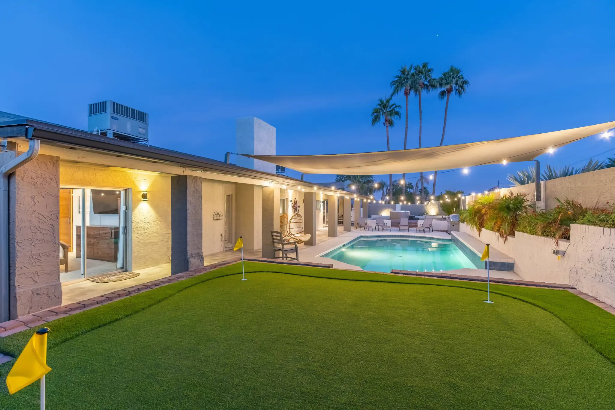A landscaped vacation home backyard at dusk features a putting green, an illuminated pool, and a covered patio. String lights draped over a sailcloth create a warm, inviting glow.