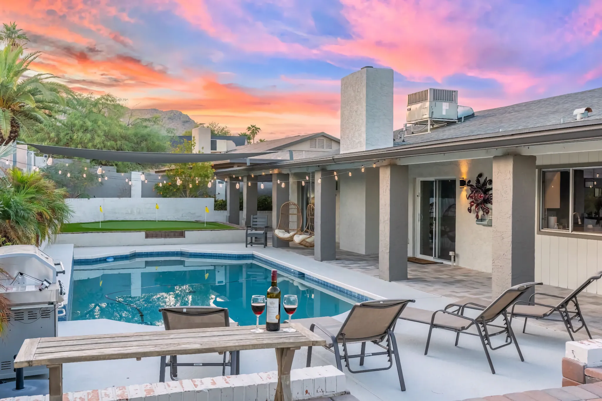 A luxurious patio and pool area at sunset, featuring lounge chairs, a grill, a putting green, and a picnic table with wine and glasses.
