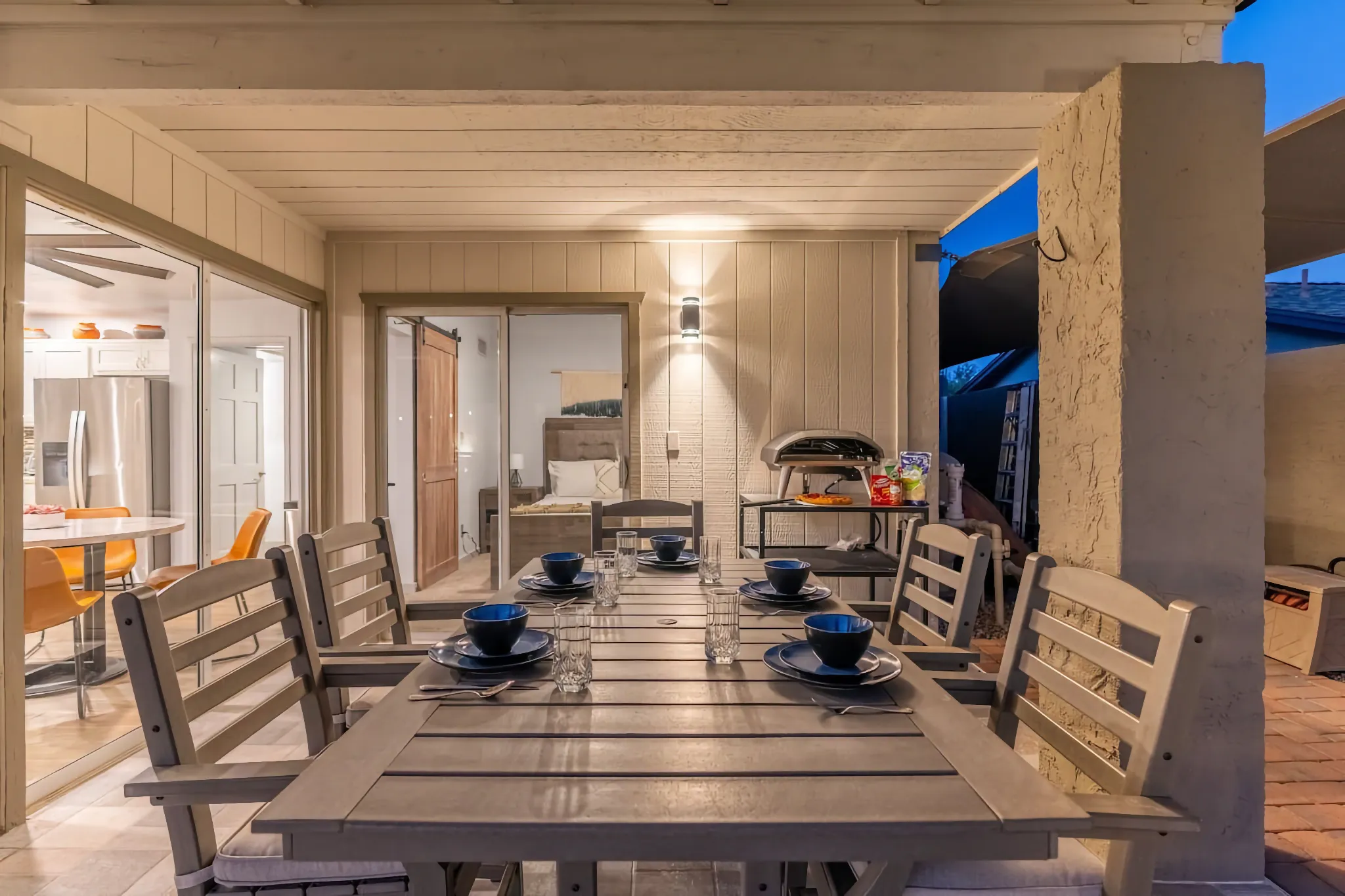 A covered outdoor patio features a slatted gray dining table set for five with blue dishware. Sliding glass doors open to show the indoor kitchen and bedroom beyond.