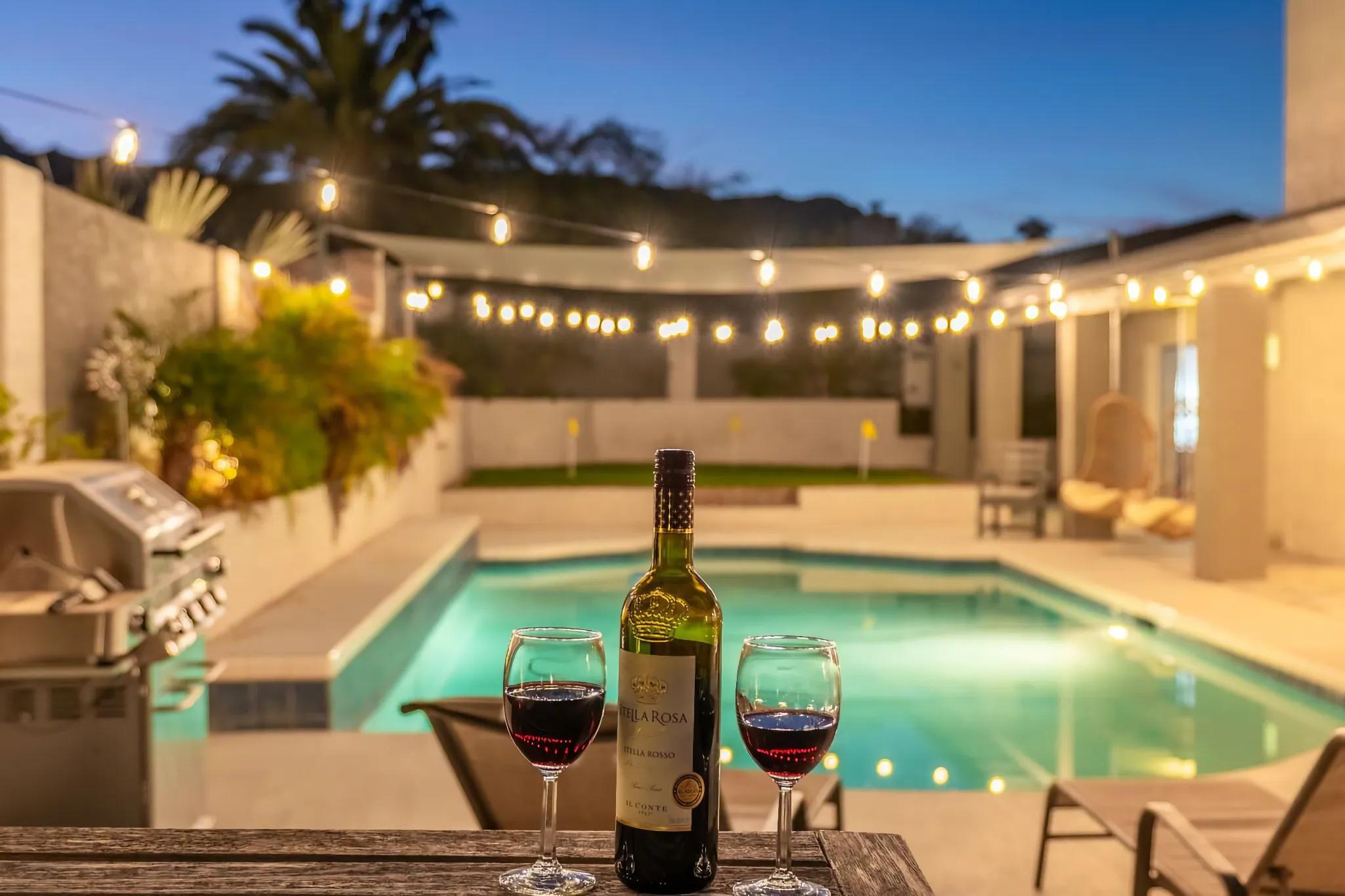 A bottle and two glasses of red wine are centered on a wooden table, with an illuminated backyard swimming pool and warm string lights visible in the background at night.