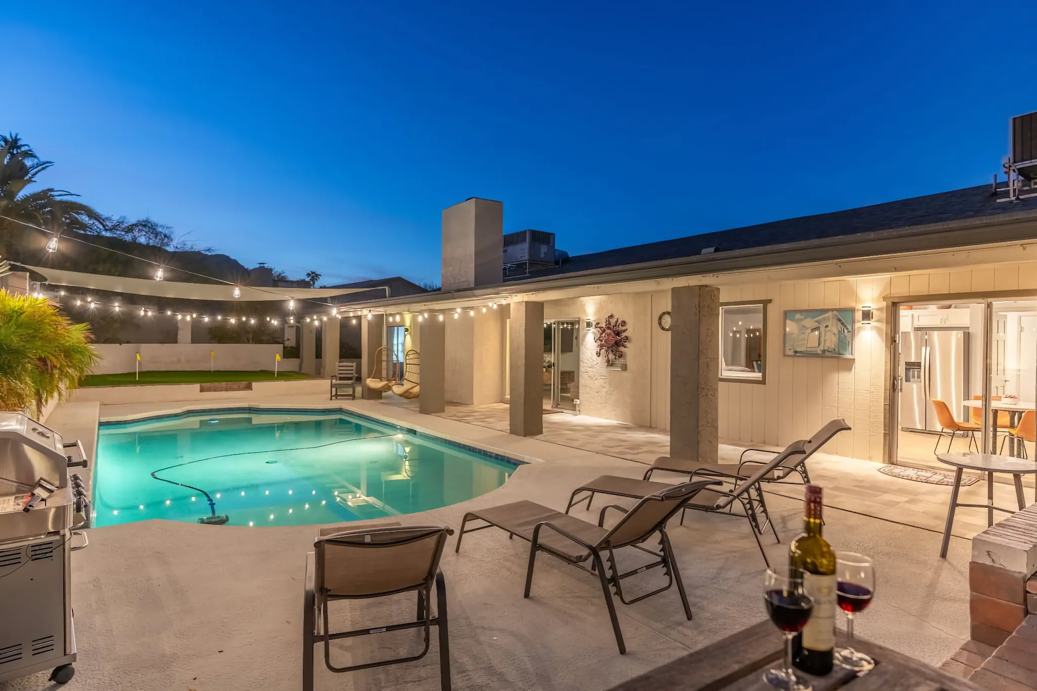 A backyard oasis at twilight with an illuminated pool and lounge chairs. String lights span the patio area, and a bottle of wine rests on a table, creating an inviting evening scene.