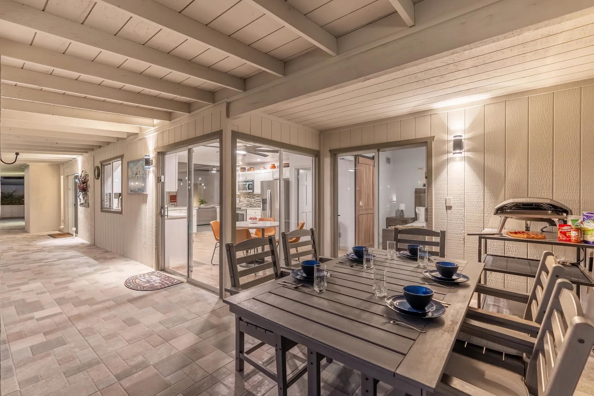 A covered outdoor patio featuring a wooden dining table set for five with blue place settings. Sliding glass doors provide a view of the brightly lit interior kitchen and bedroom.
