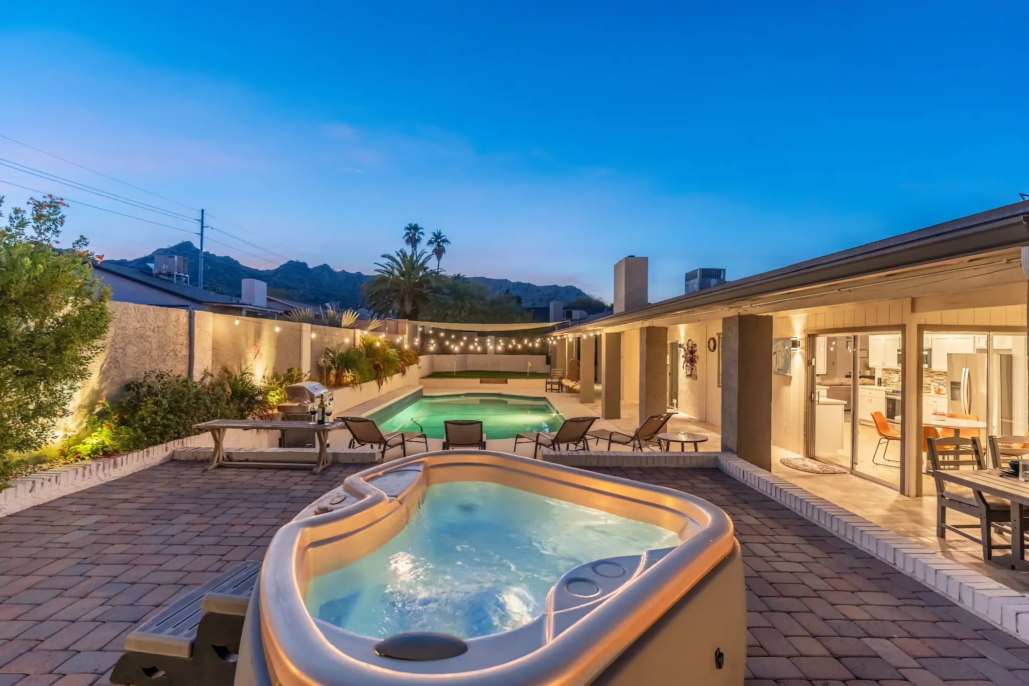 A luxurious backyard at twilight with a bubbling hot tub and a lighted swimming pool, connected to the house by a paved patio under string lights.