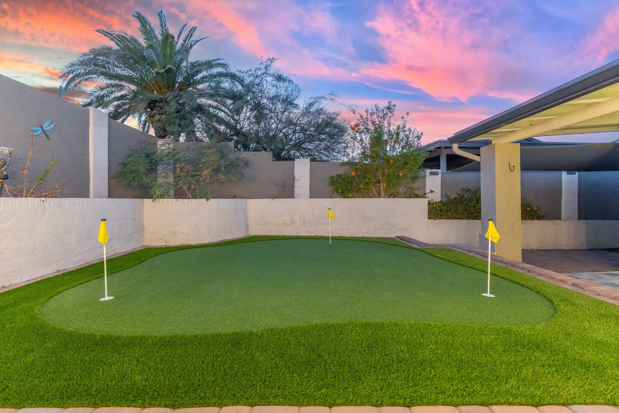 An immaculate backyard putting green featuring three yellow-flagged holes, set against high privacy walls and a colorful sunset.