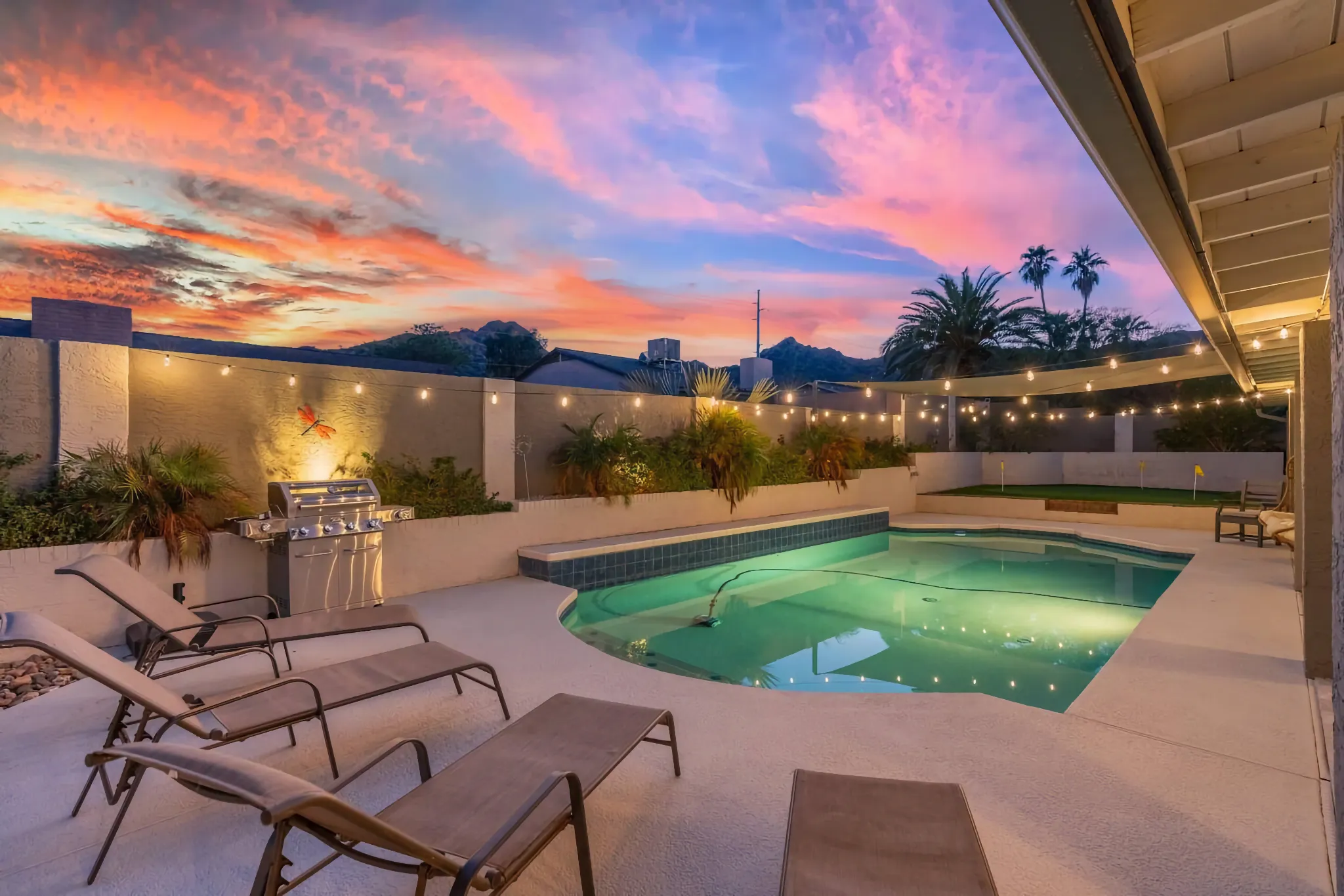 An illuminated backyard swimming pool area at dusk. A dramatic sunset features orange and pink clouds over mountains. The deck holds lounge chairs, a stainless steel grill, and bistro lights.
