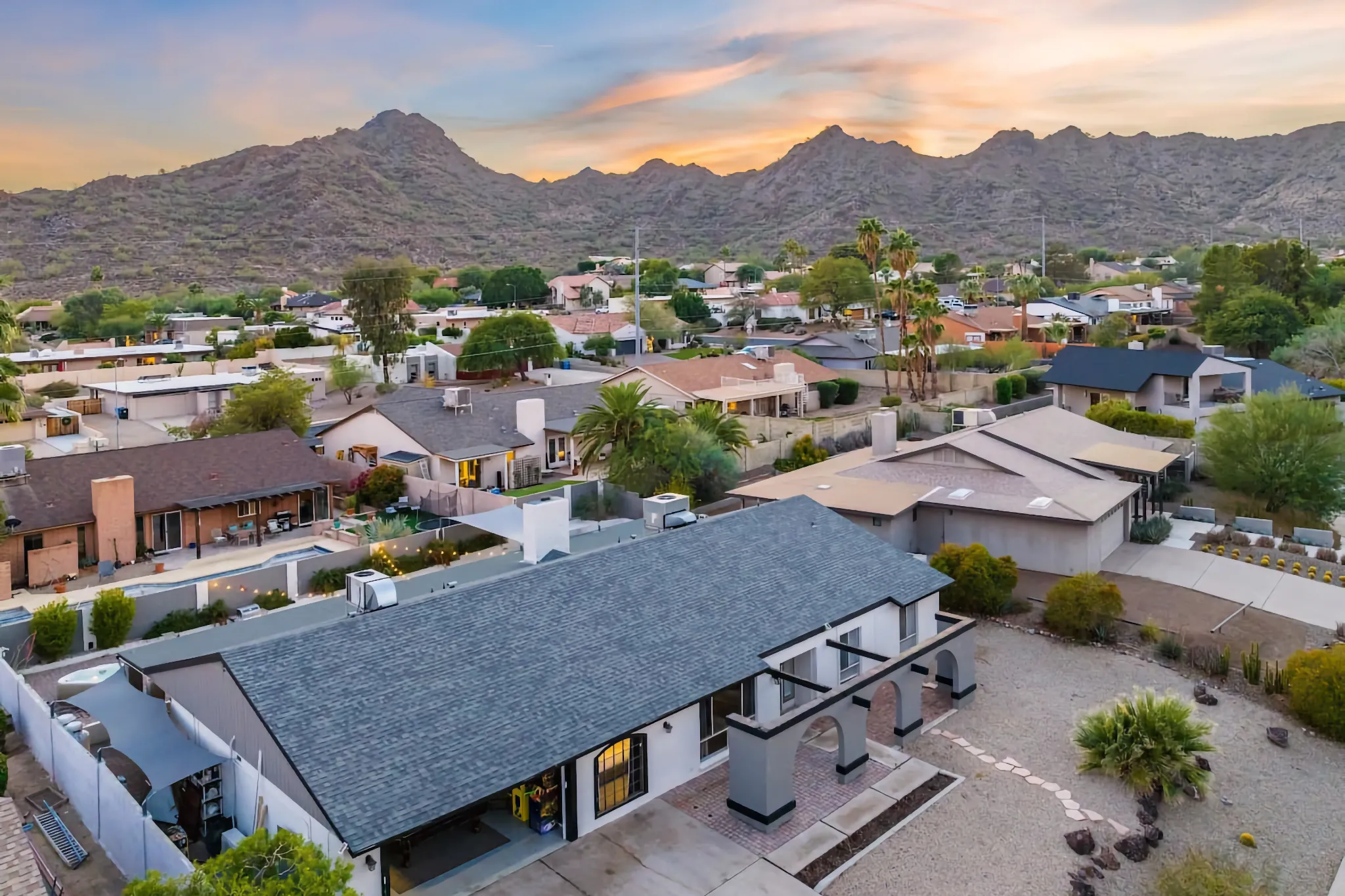 An aerial view of a suburban neighborhood and homes set against rugged desert mountains under a dramatic orange and pink sunset sky.