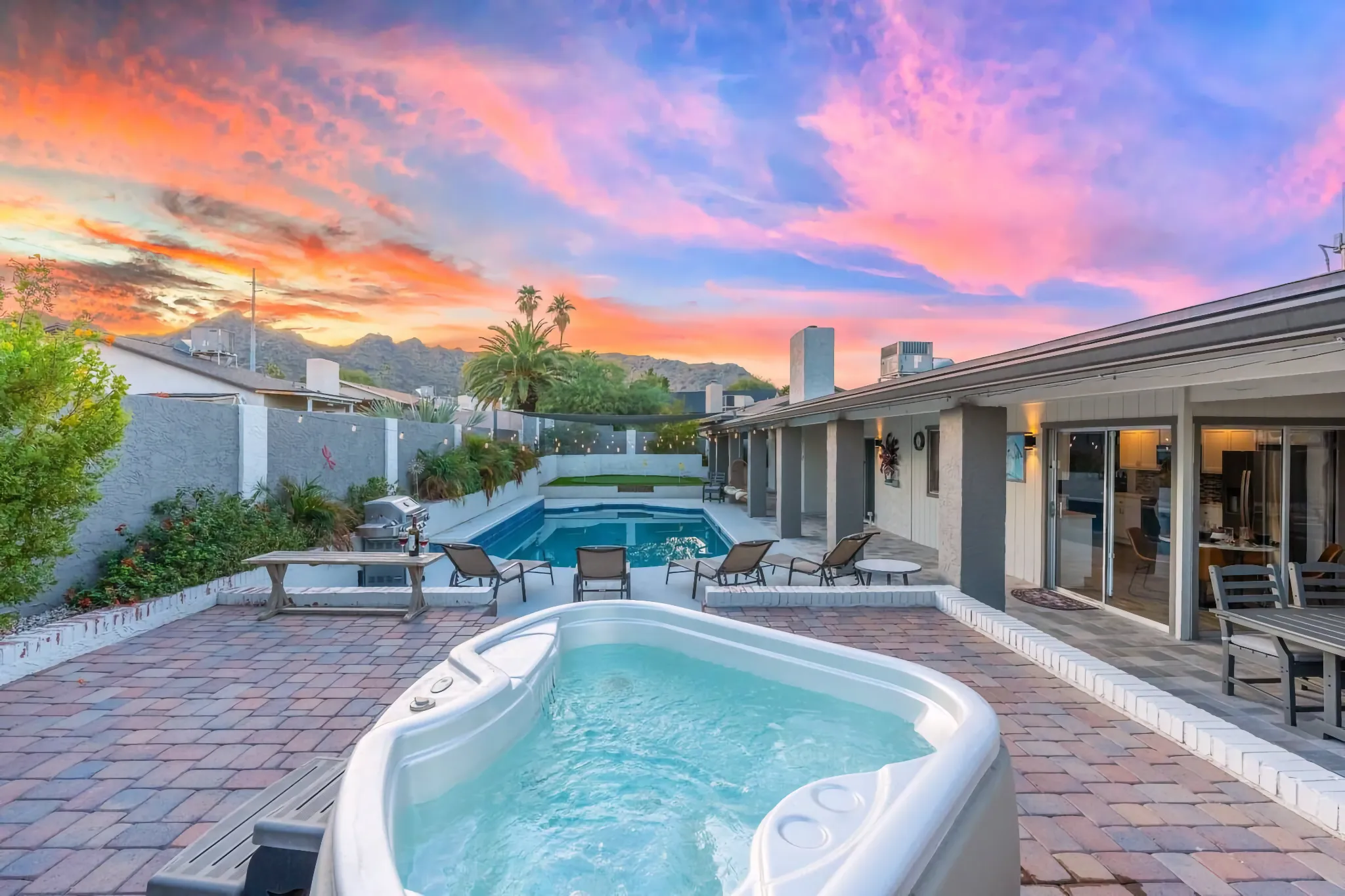 A residential backyard with a hot tub and swimming pool, surrounded by lounge chairs and pavers, under a vivid pink and orange sunset.