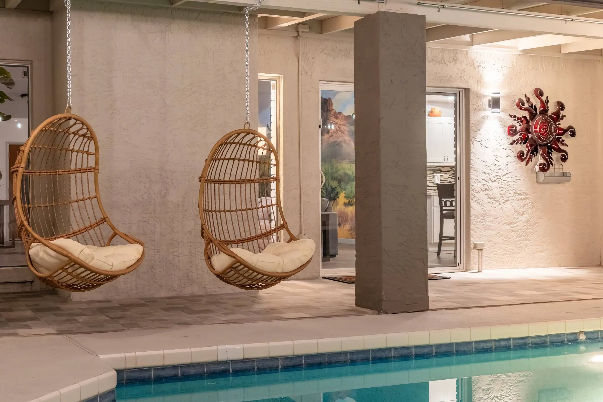 Two rattan hanging chairs with white cushions are suspended over a swimming pool edge on a covered patio lit by warm evening lights.