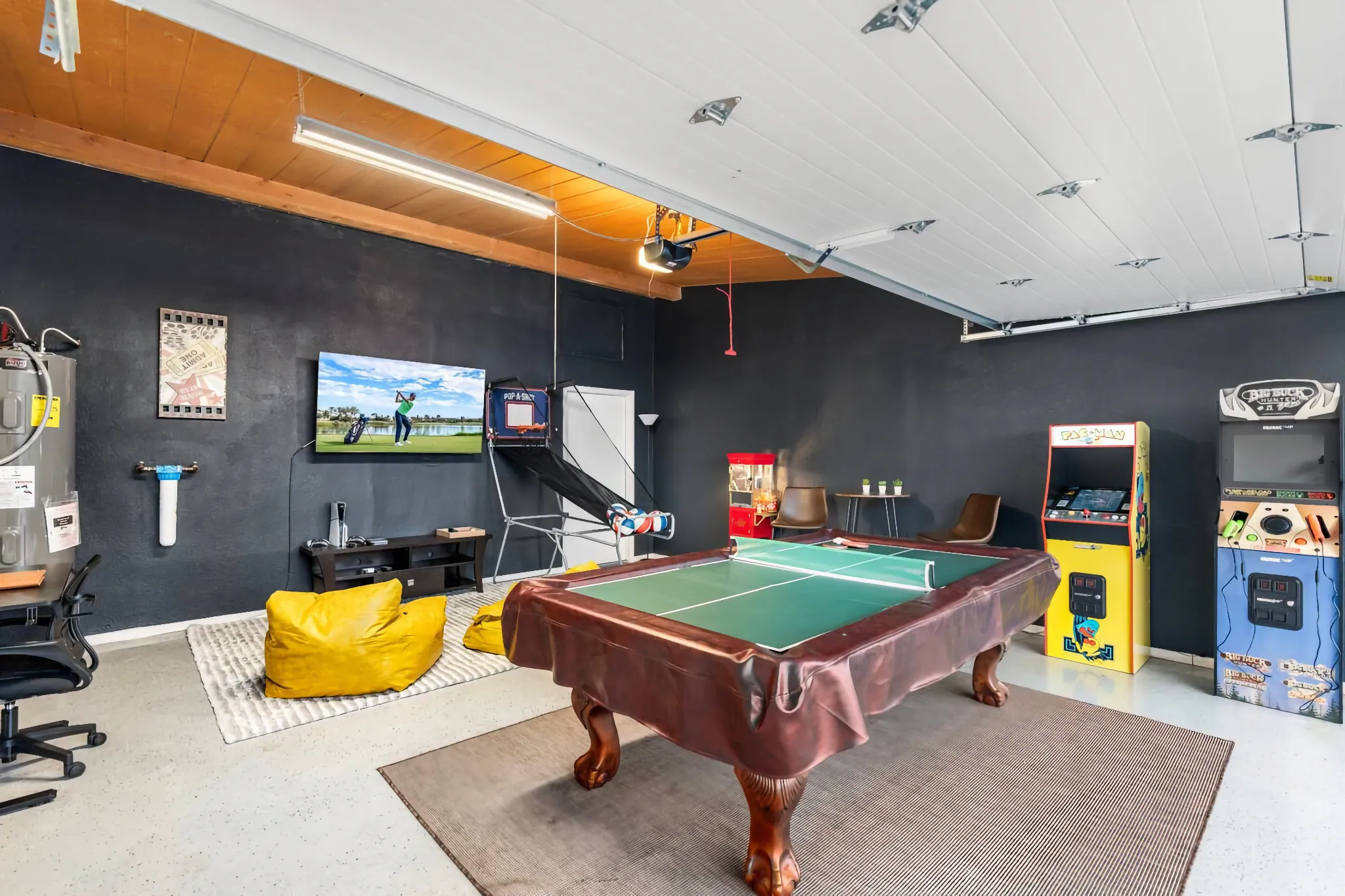 A converted garage game room featuring a covered pool table in the center, flanked by bright arcade cabinets, a large TV, and yellow bean bag chairs.