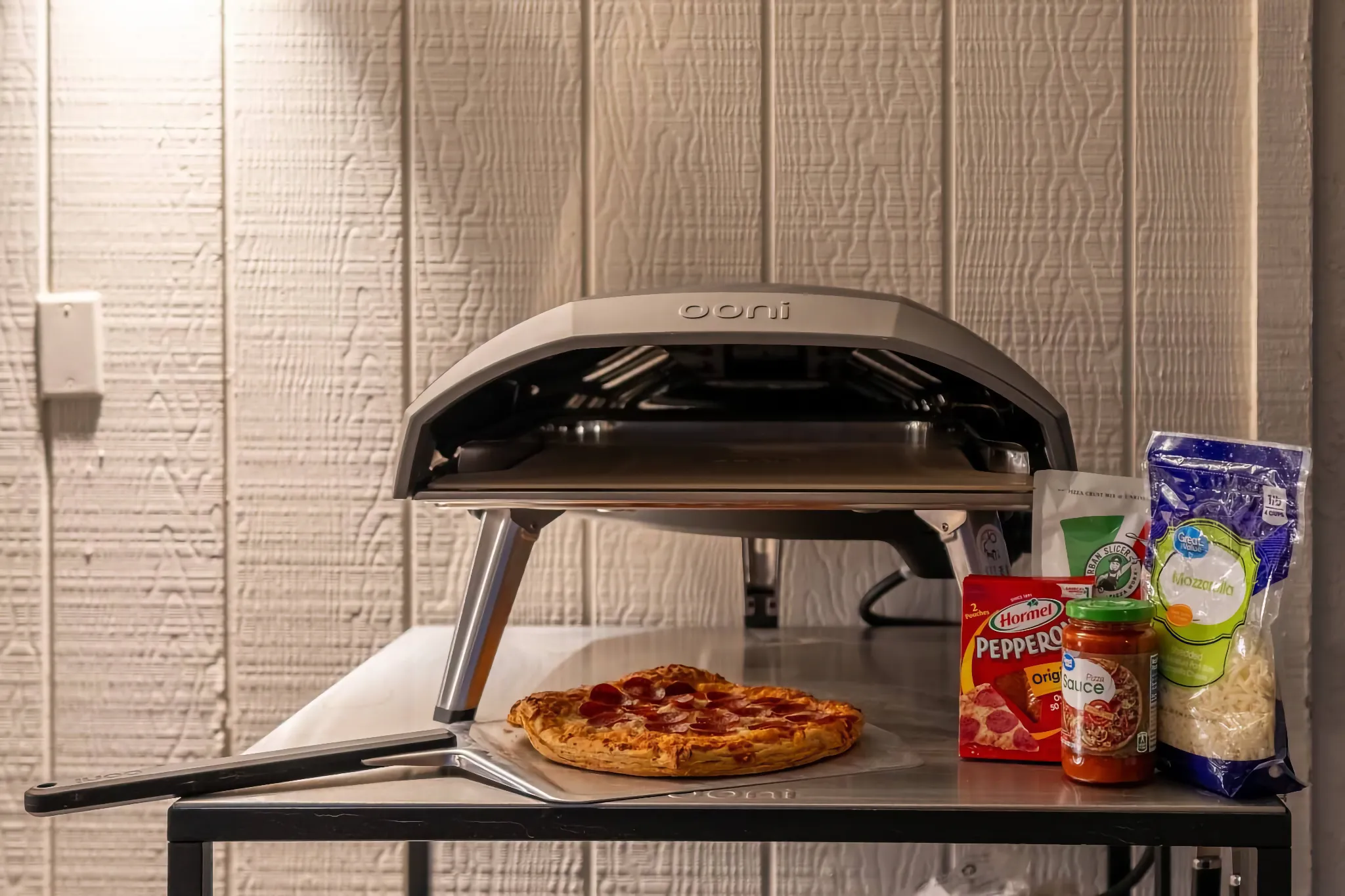 A countertop setup featuring a gray Ooni pizza oven, a fresh pepperoni pizza on a peel, and ingredients like sauce, cheese, and pepperoni.