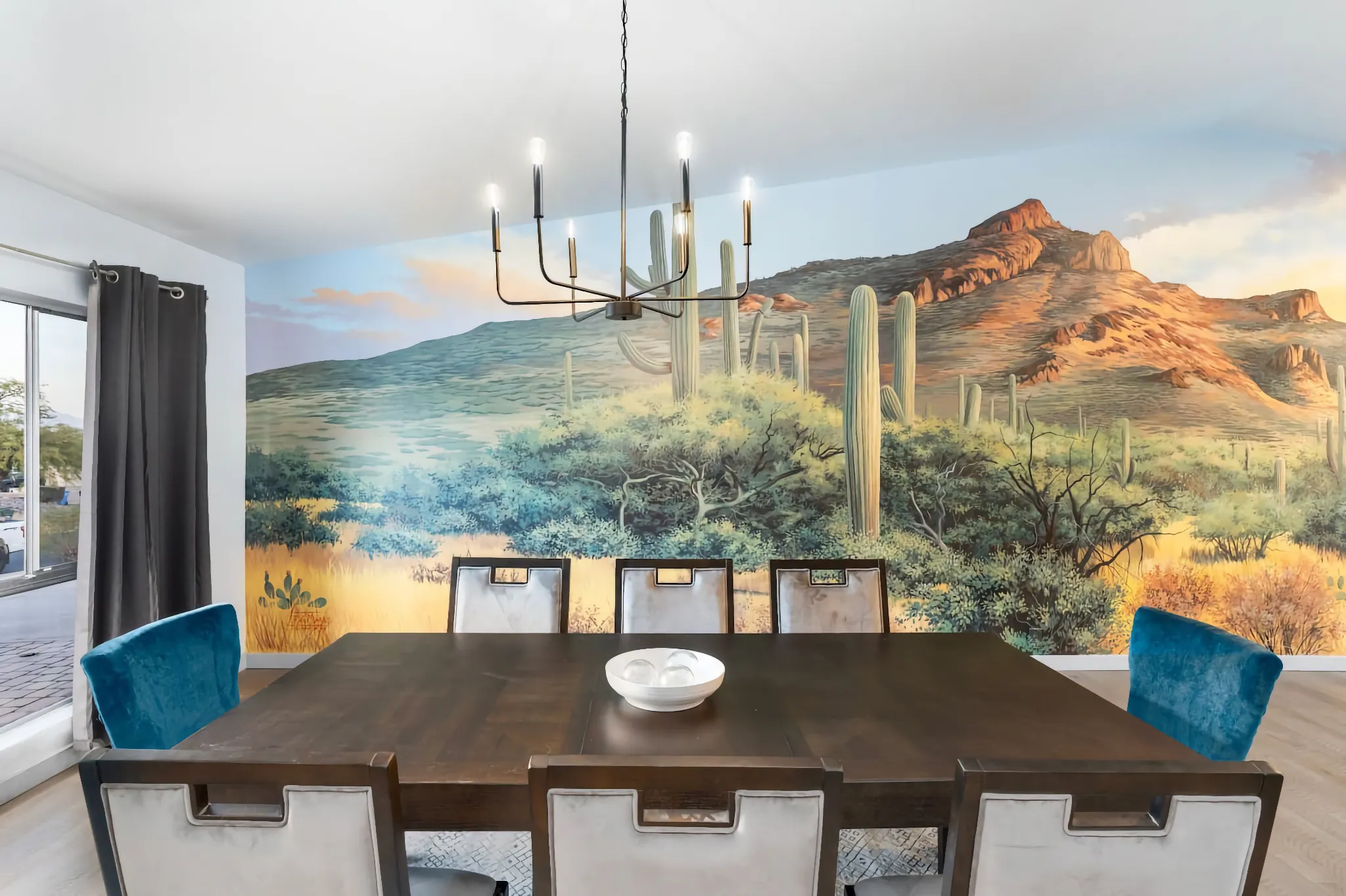 A modern dining room featuring a dark wood table and gray and turquoise chairs, set against a vibrant wall mural of a desert scene with saguaro cacti and mountains.