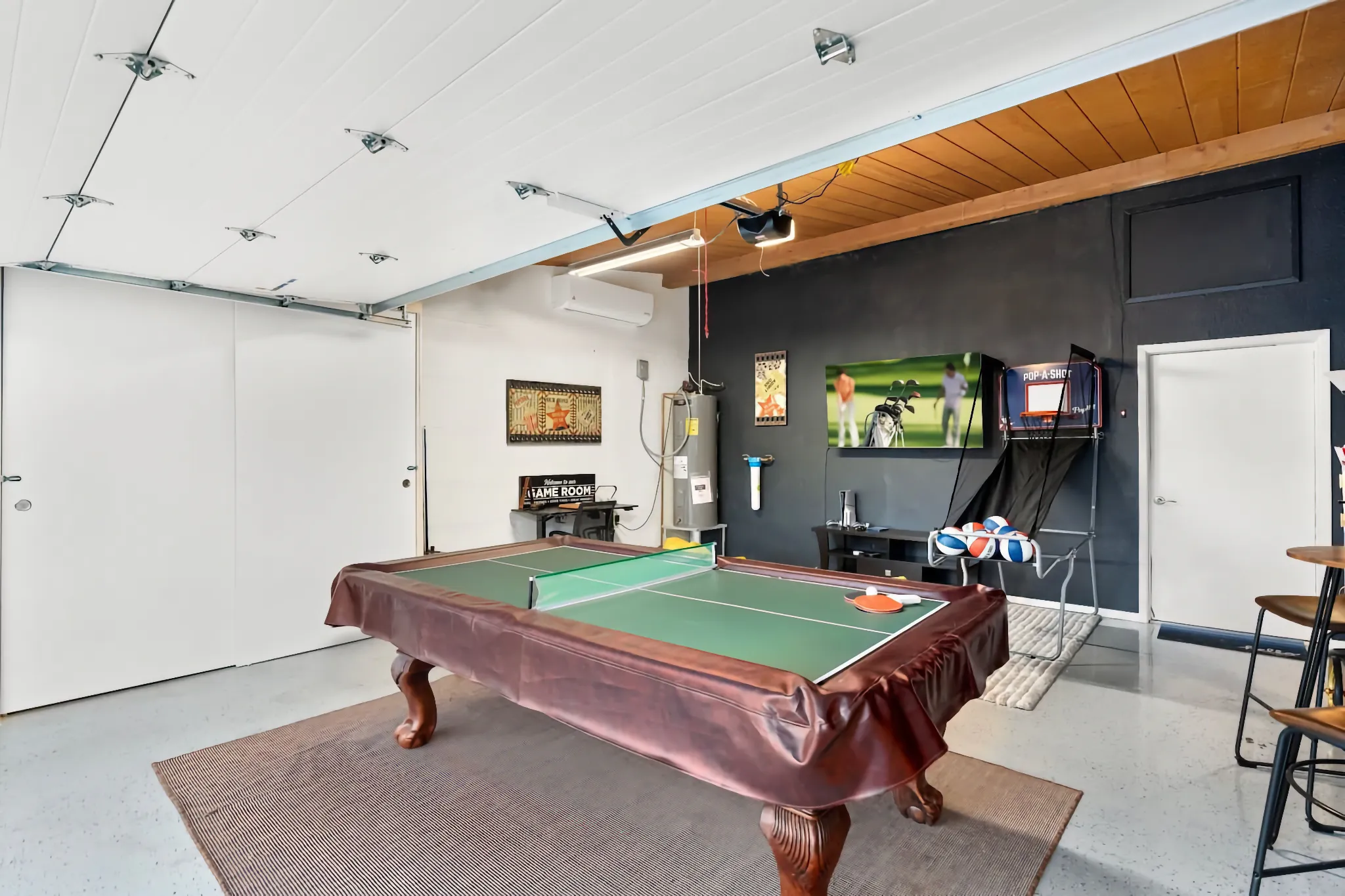 A converted garage space now serves as a game room, featuring a pool table with a ping-pong top. The room includes a TV, a water heater, a basketball arcade game, and bar seating along a black accent wall.