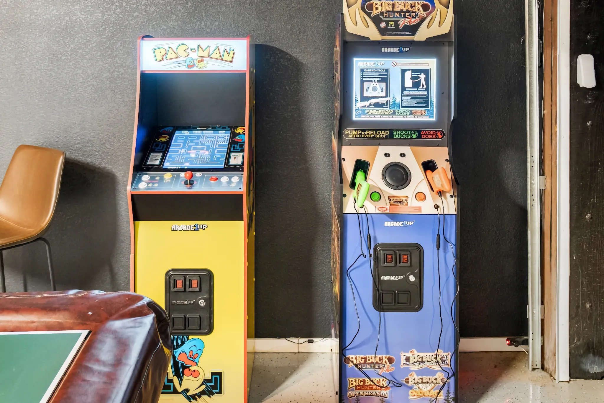 Two standing arcade cabinets, Pac-Man (yellow) and Big Buck Hunter (blue), are ready for use in a recreation area. A brown upholstered chair is visible on the left.