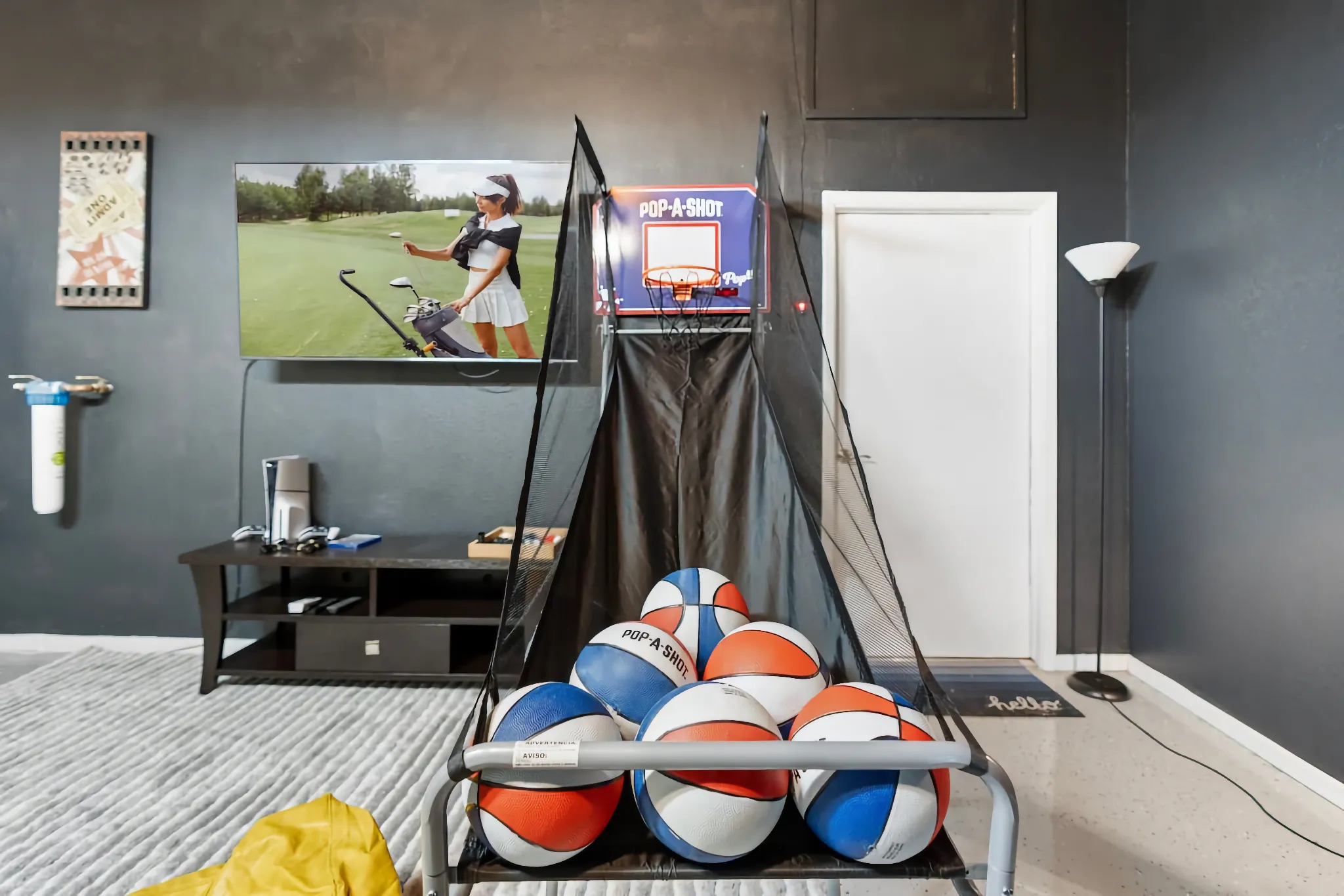A game room with dark gray walls includes a basketball Pop-A-Shot arcade game. In the background, a TV displays a video of a female golfer.