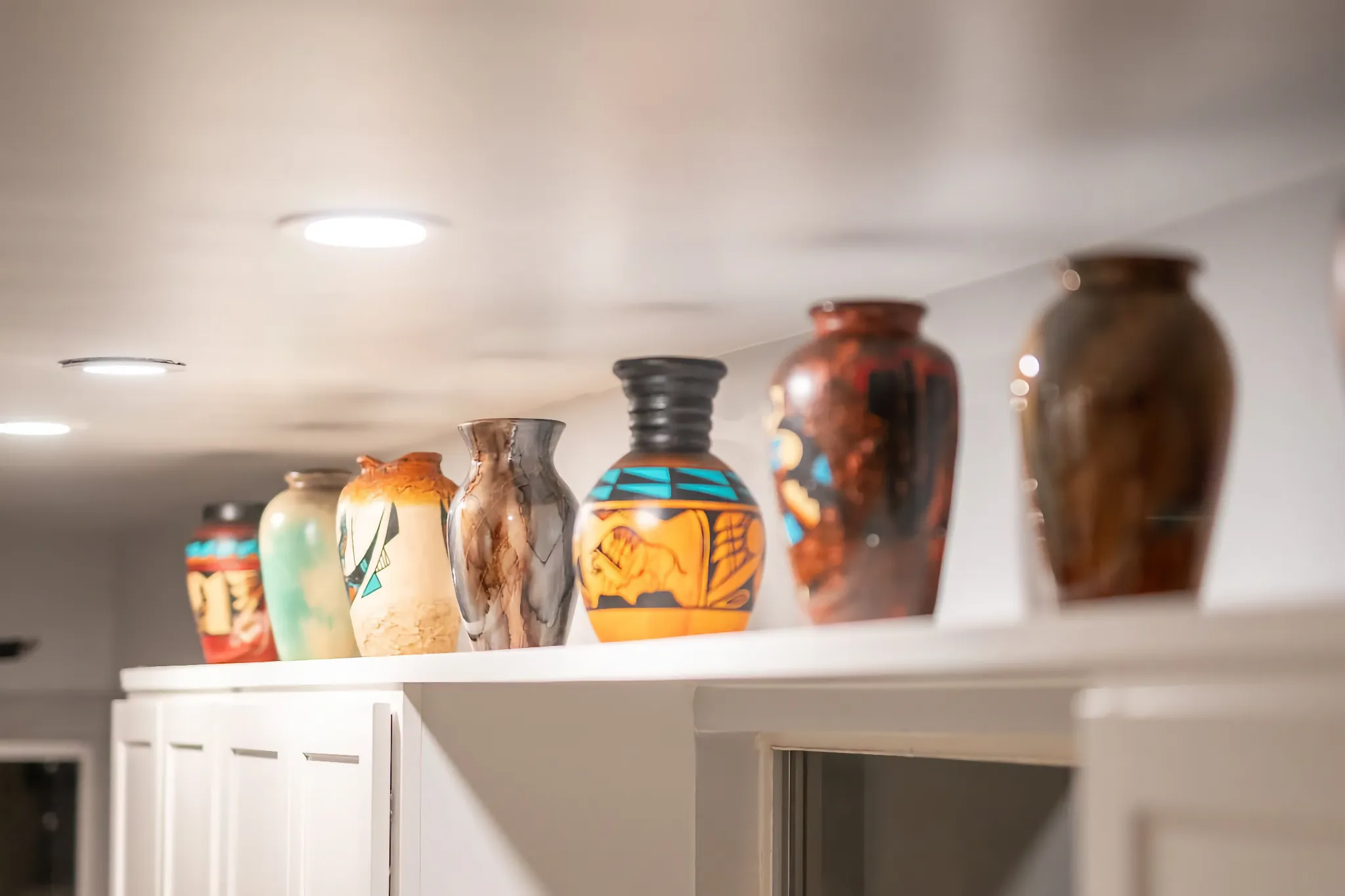 A line of colorful, decorative Southwestern-style vases sits on top of white cabinets in a well-lit interior room with recessed lighting.