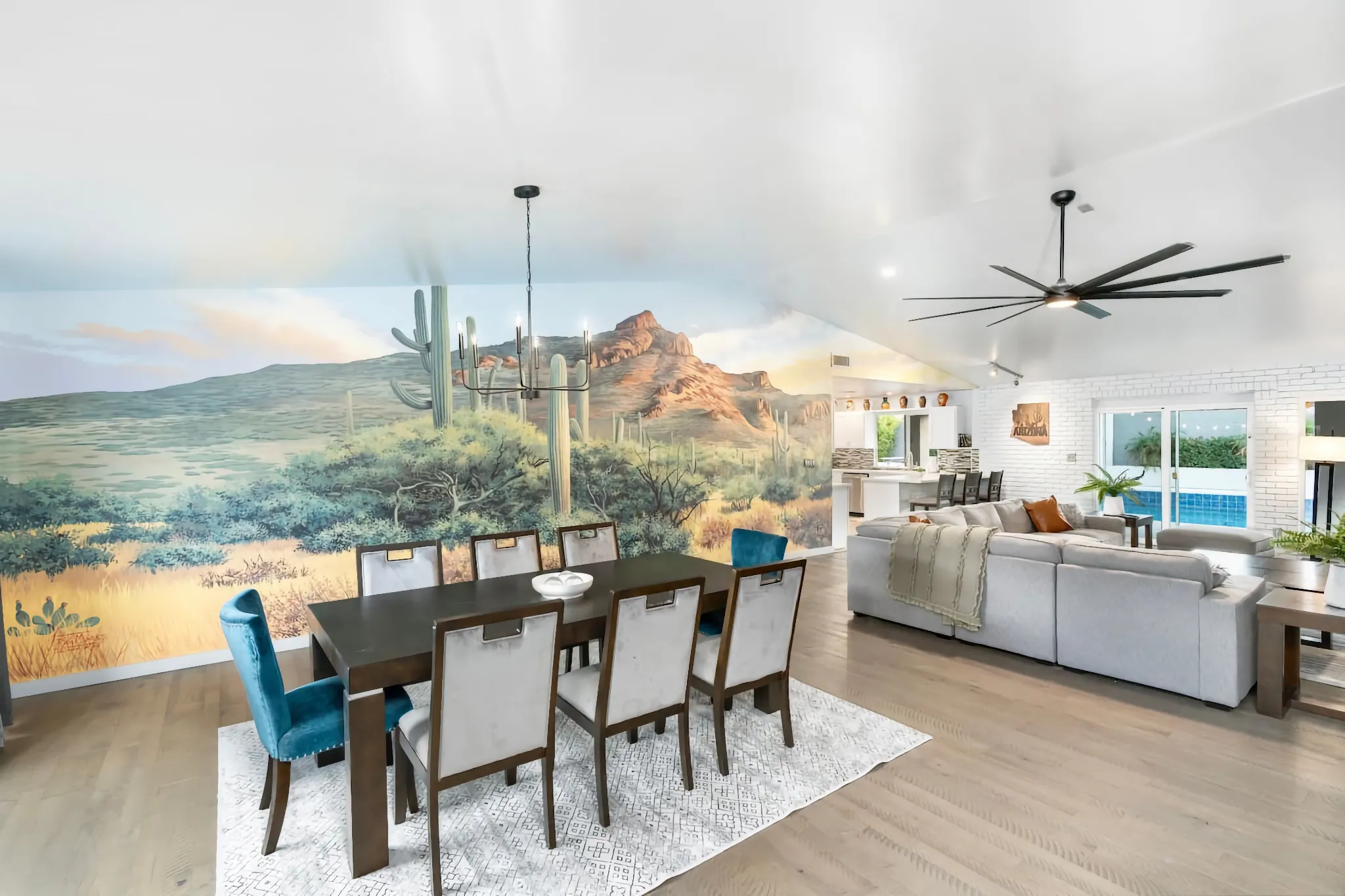 A bright, open-concept room with a desert mural covering one wall. A dark dining table with velvet chairs sits on a rug near a large gray sectional sofa.