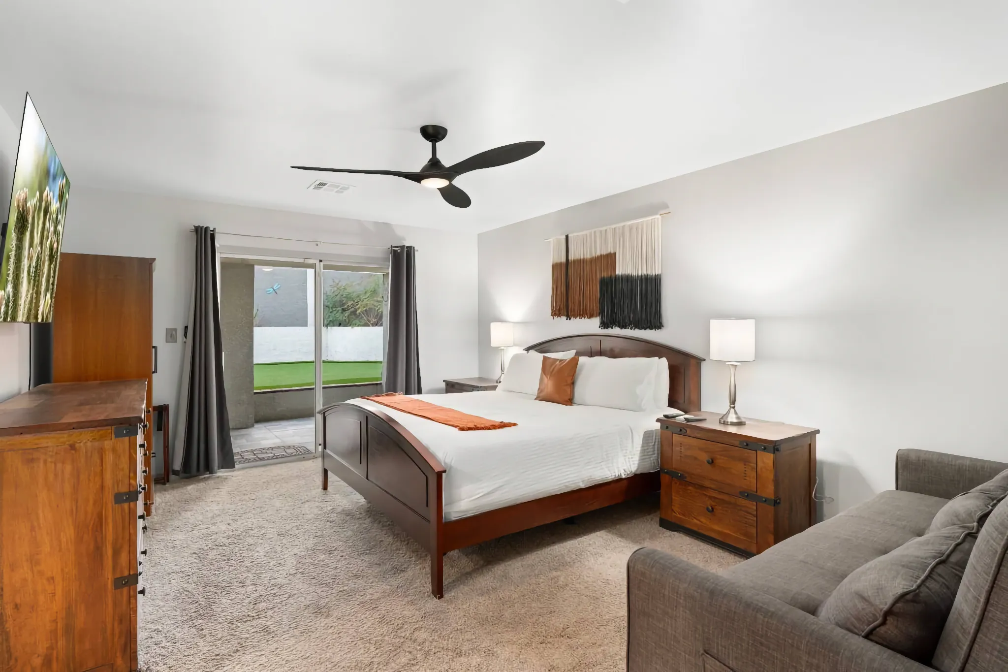 A modern bedroom with a dark wood bed, matching furniture, and a gray sofa. Sliding doors lead to a patio with green turf.