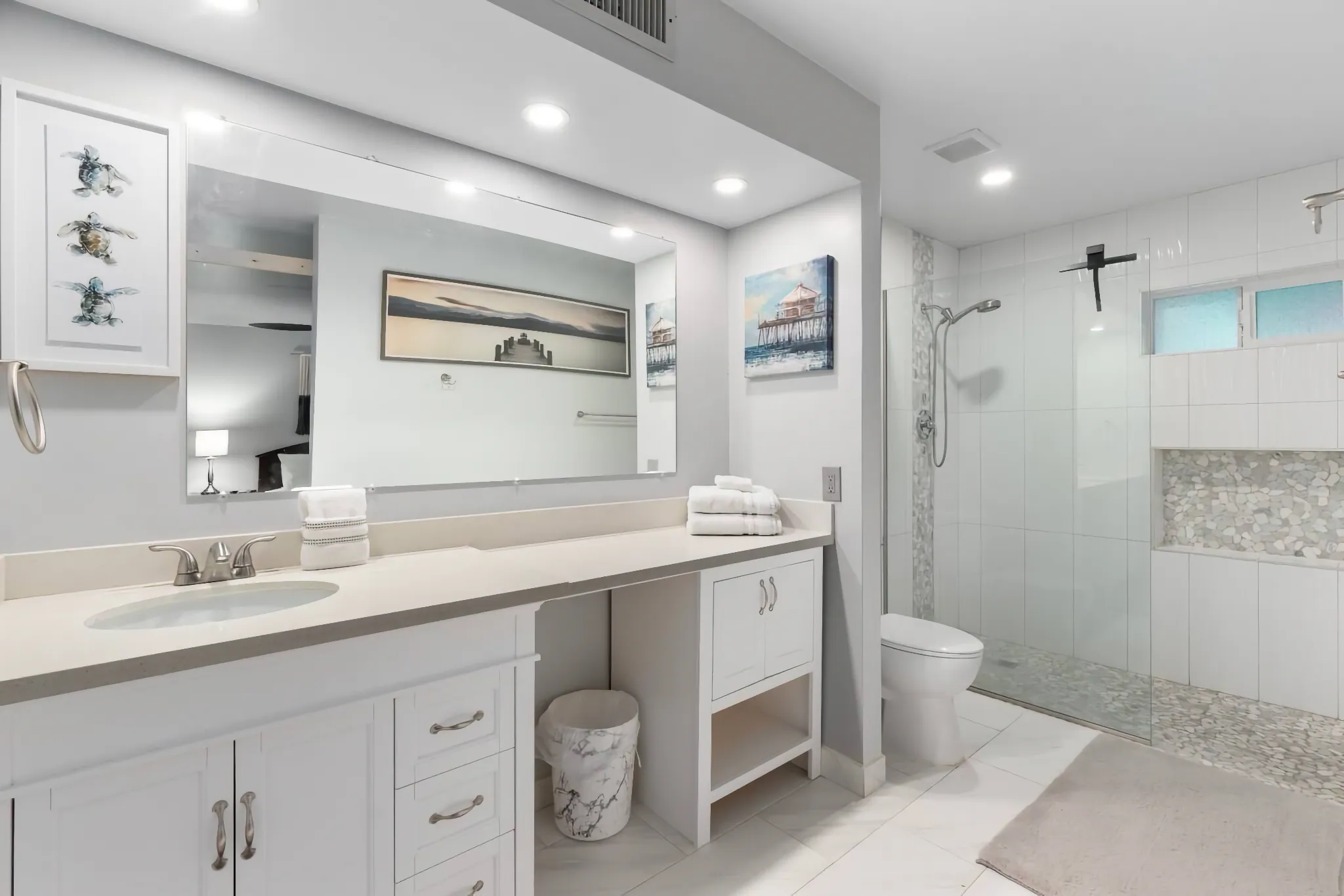 A clean, modern bathroom with a white cabinet vanity and a large mirror over a light gray counter. A glass shower enclosure features white tile walls and pebble stone accents.
