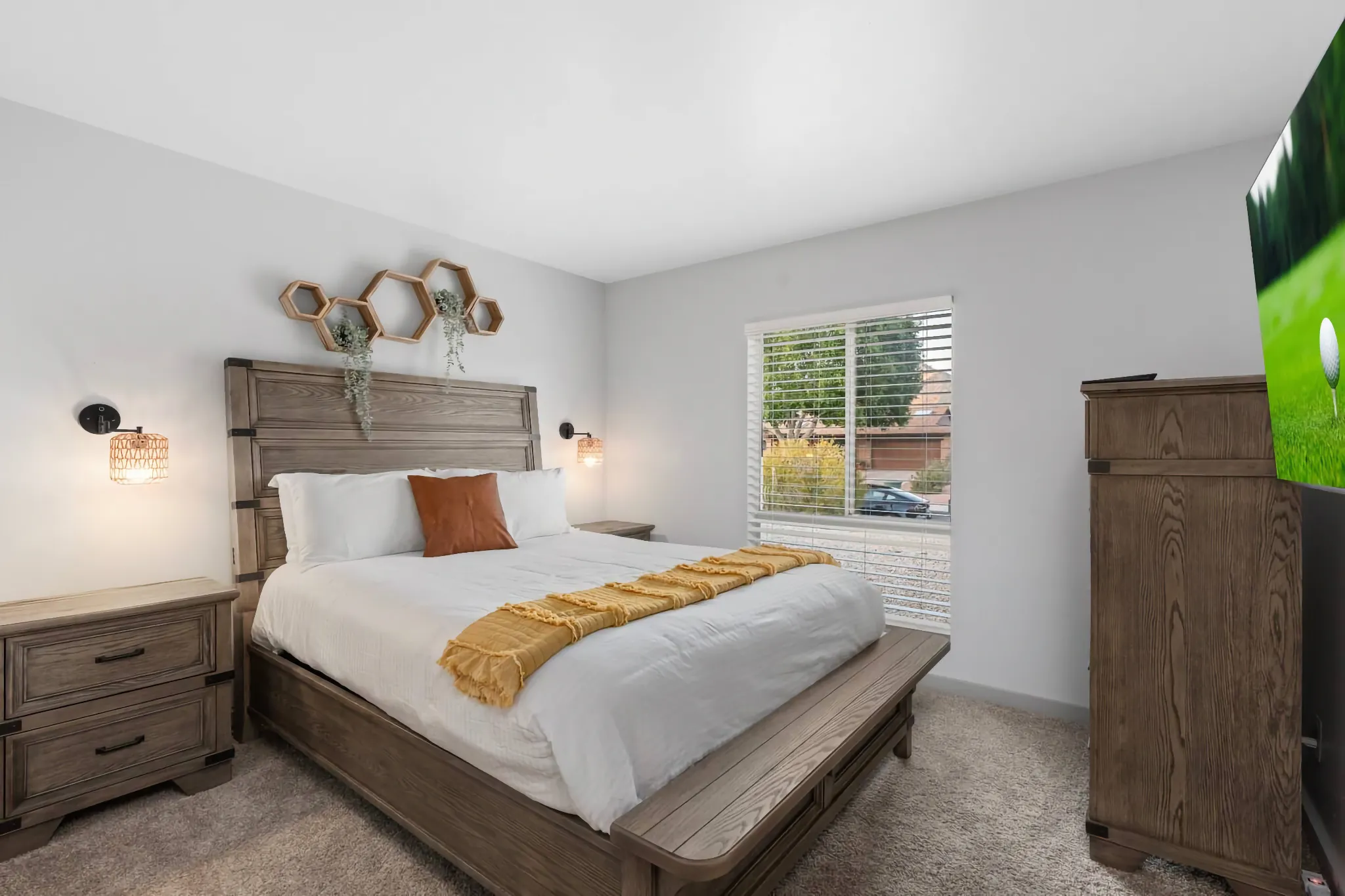 A modern bedroom with a rustic wood bed frame, white bedding, and a yellow accent blanket. Hexagonal shelves and decorative lights flank the headboard, with matching wood nightstands and dresser.