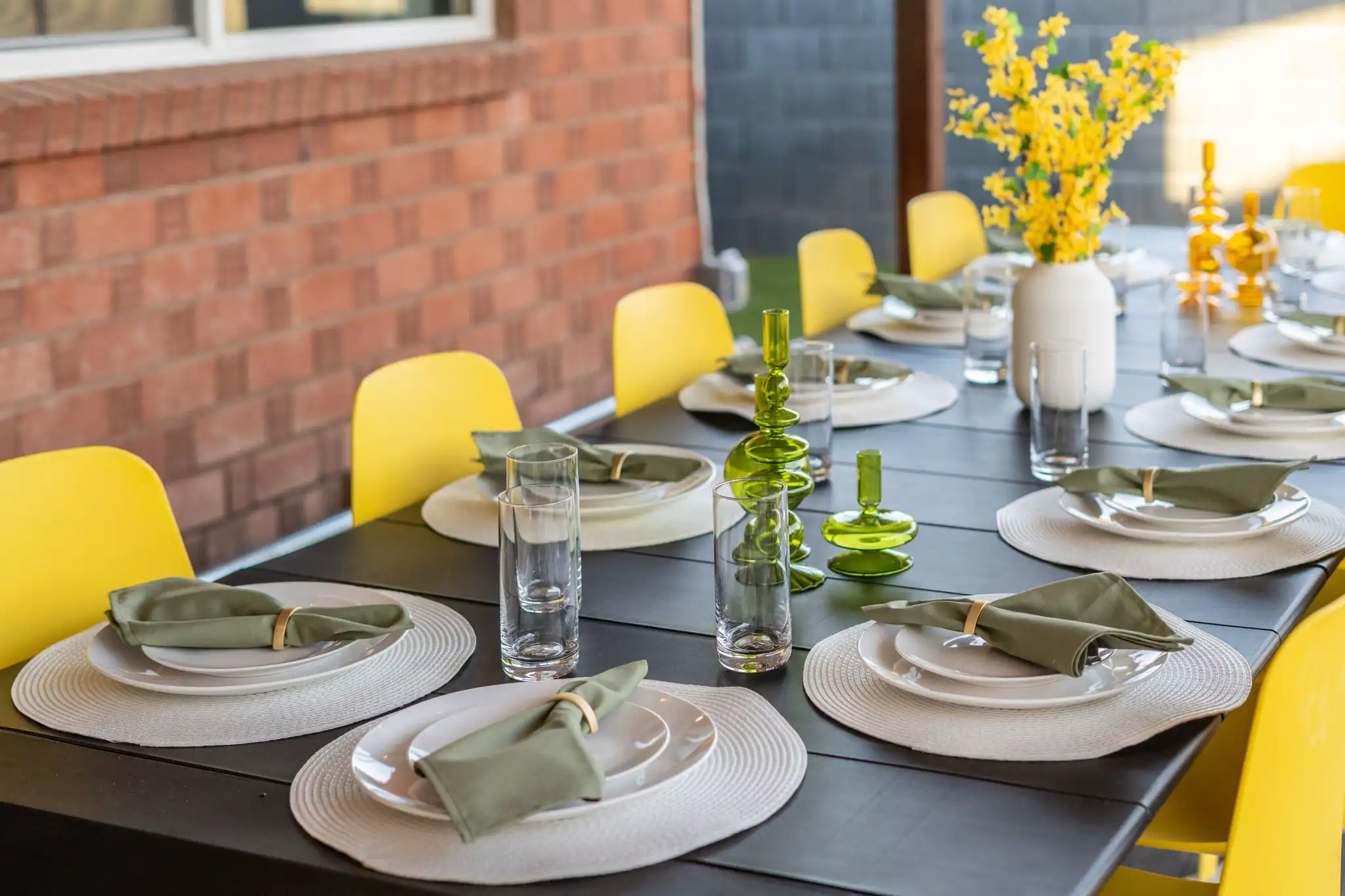 A dark outdoor patio table is fully set with bright yellow chairs, white plates, green napkins, and vibrant yellow floral arrangements for dining.