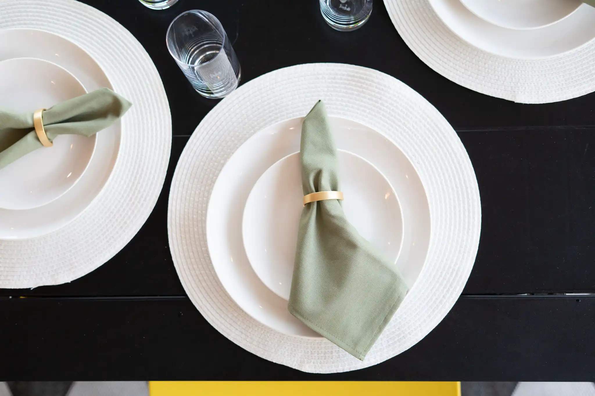 A clean table setting featuring a white placemat and plates on a black table. A sage green napkin is folded and held by a gold napkin ring.