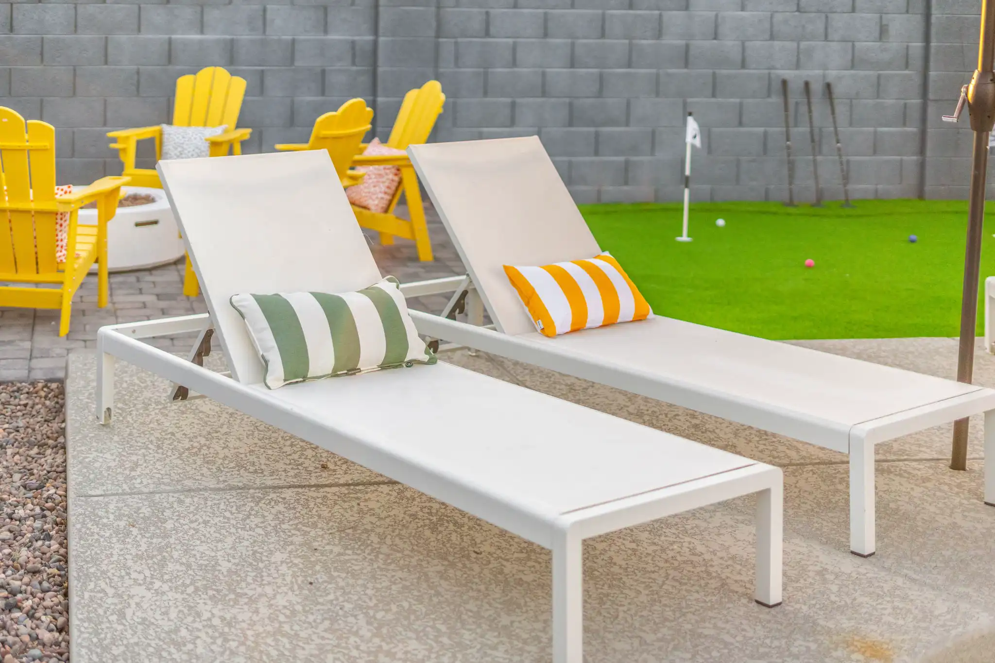 Two modern white lounge chairs with striped pillows are placed on a patio. The background shows yellow Adirondack chairs near a fire pit and a synthetic turf putting green.