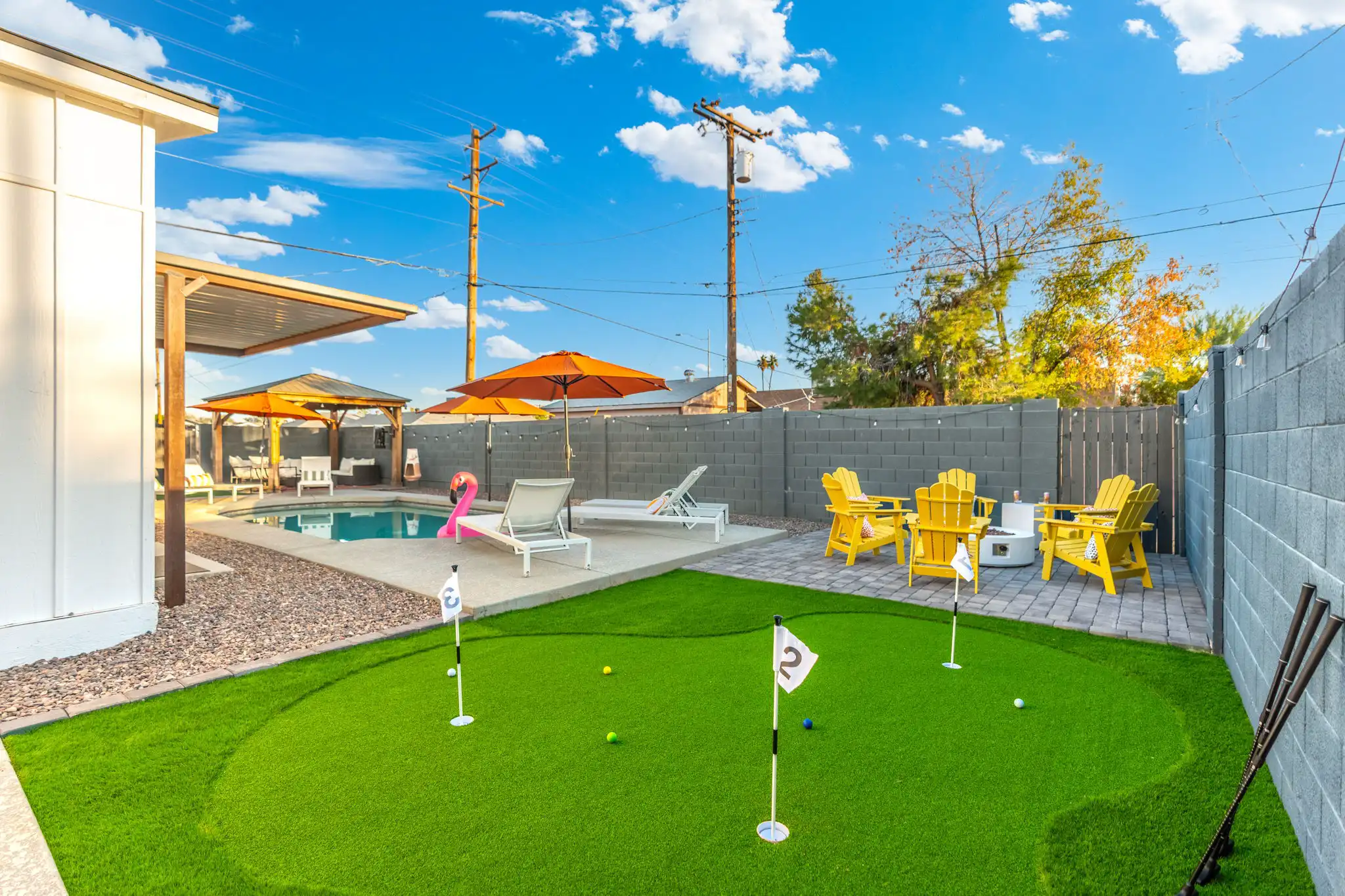 A sunny backyard offering a swimming pool with lounge chairs, shaded seating under a pergola, and a curved putting green in the foreground.