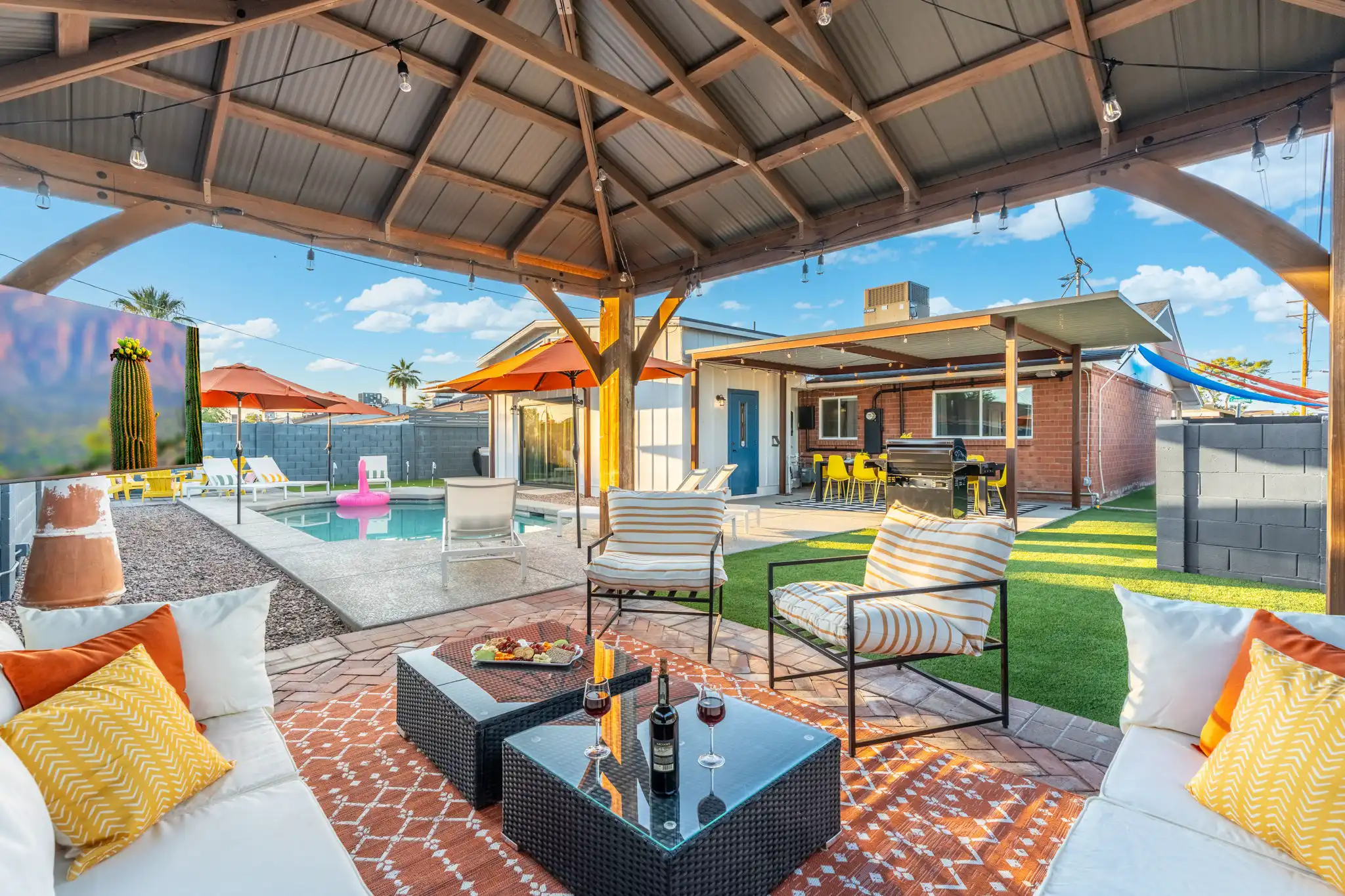 A bright, covered outdoor patio features comfortable white furniture and two coffee tables with a bottle of wine and snacks. The space overlooks a pool area with lounge chairs and umbrellas under a sunny sky.