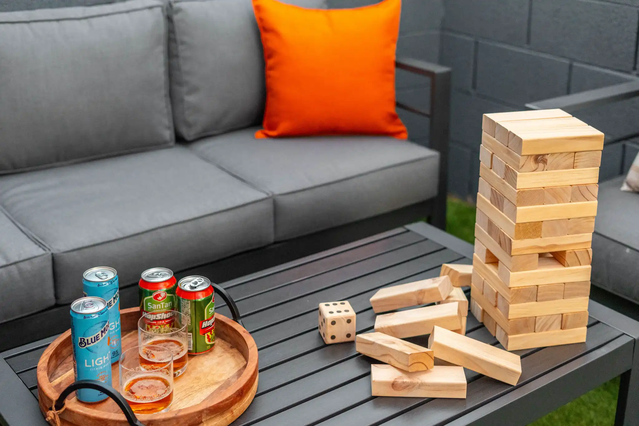 A tall wooden stacking block game and scattered pieces sit on a dark patio table, near a tray holding four beer cans and two filled glasses. A gray outdoor sofa is nearby.