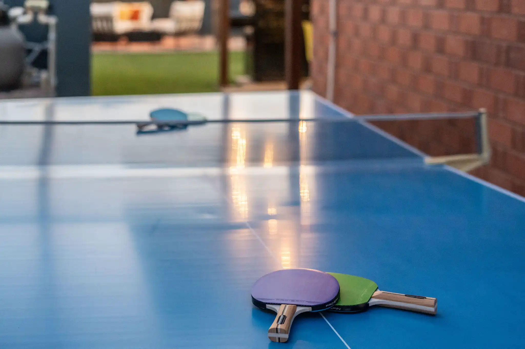 Two ping pong paddles, purple and green, rest on a blue outdoor table tennis surface, which reflects the warm sunset light.