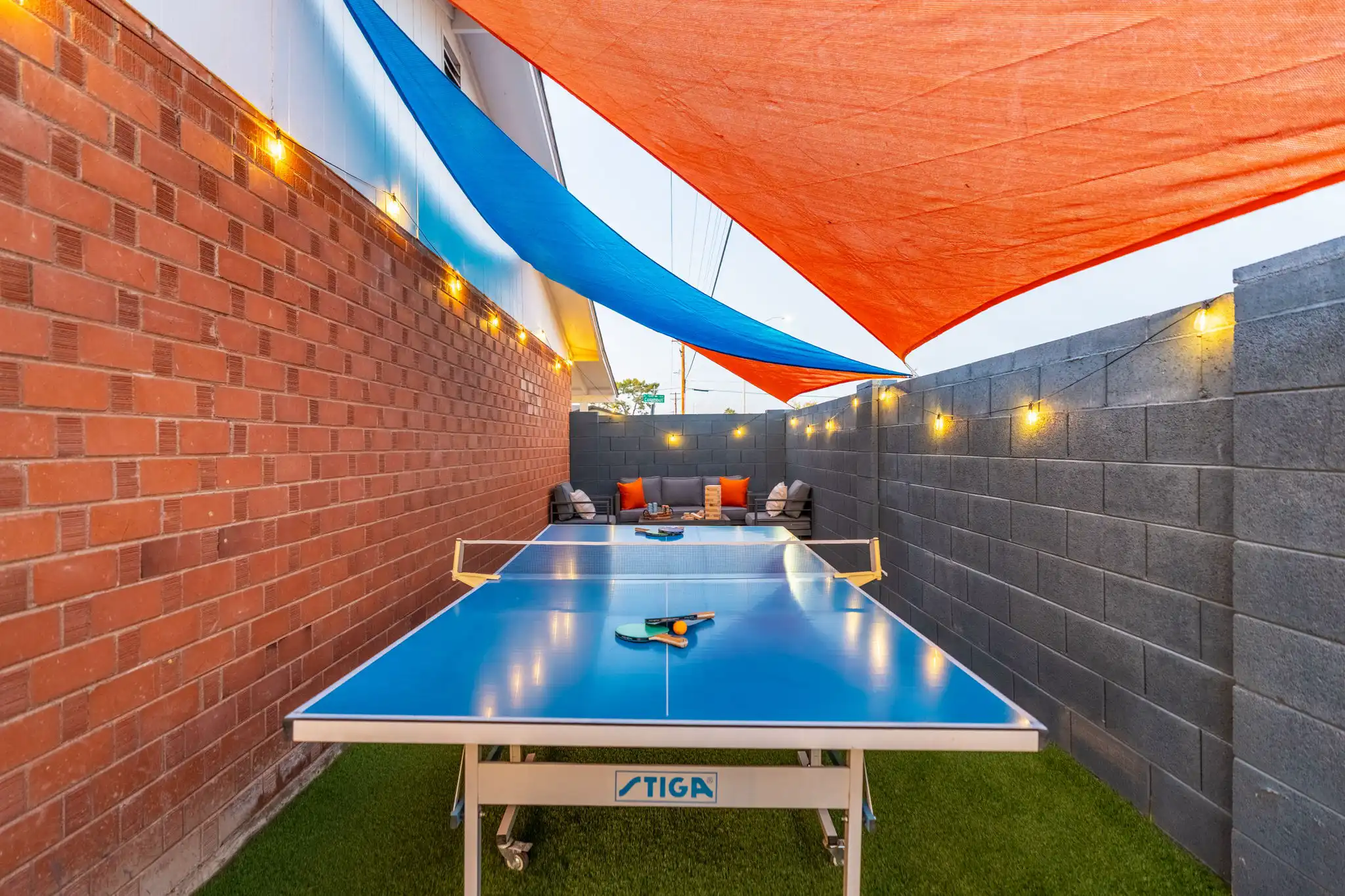 A vibrant outdoor recreation area featuring a blue ping pong table. Orange and blue shade sails hang above, illuminating the space between a red brick wall and a gray block wall.