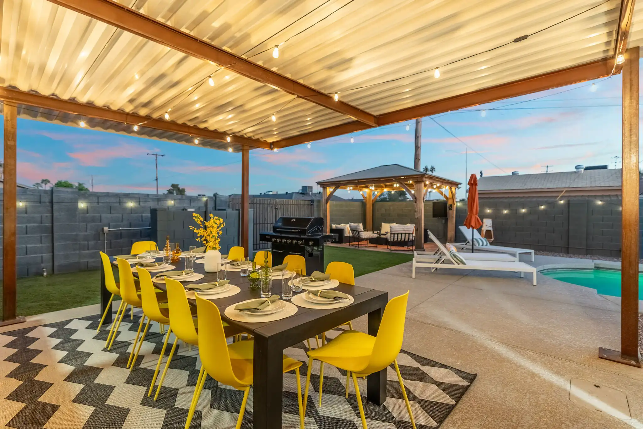 A chic, covered patio entertainment area features a long table set with ten bright yellow chairs over a geometric rug. String lights illuminate the space, which includes a grill and a seated gazebo.