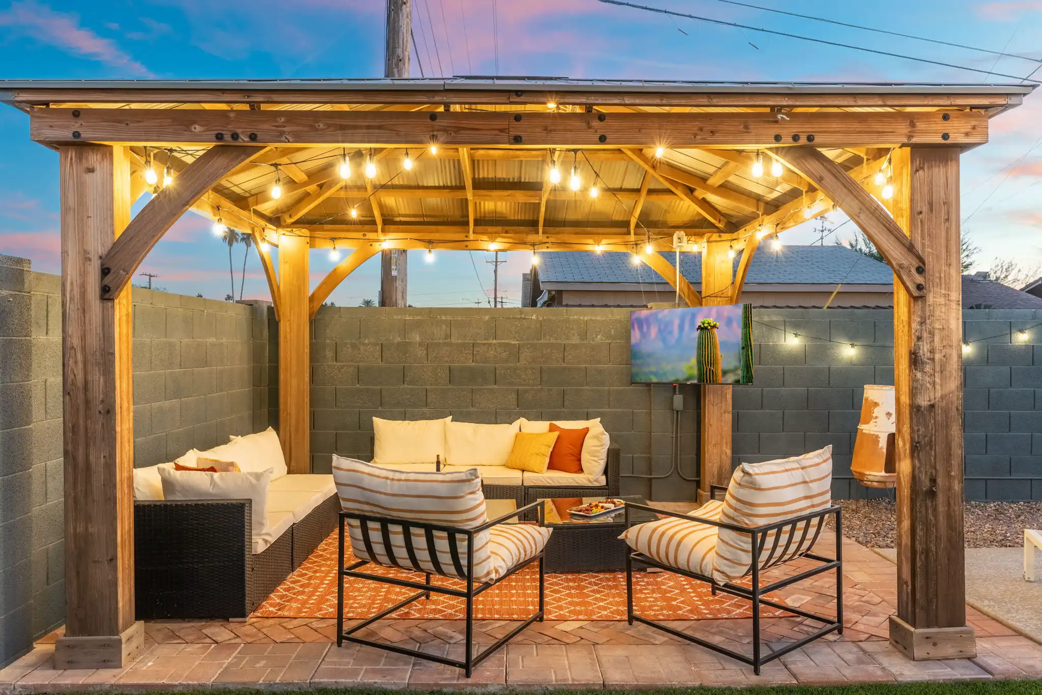 An inviting outdoor social space at a vacation rental featuring a wooden pergola, string lights, comfortable patio furniture, and a mounted TV set against a sunset sky.