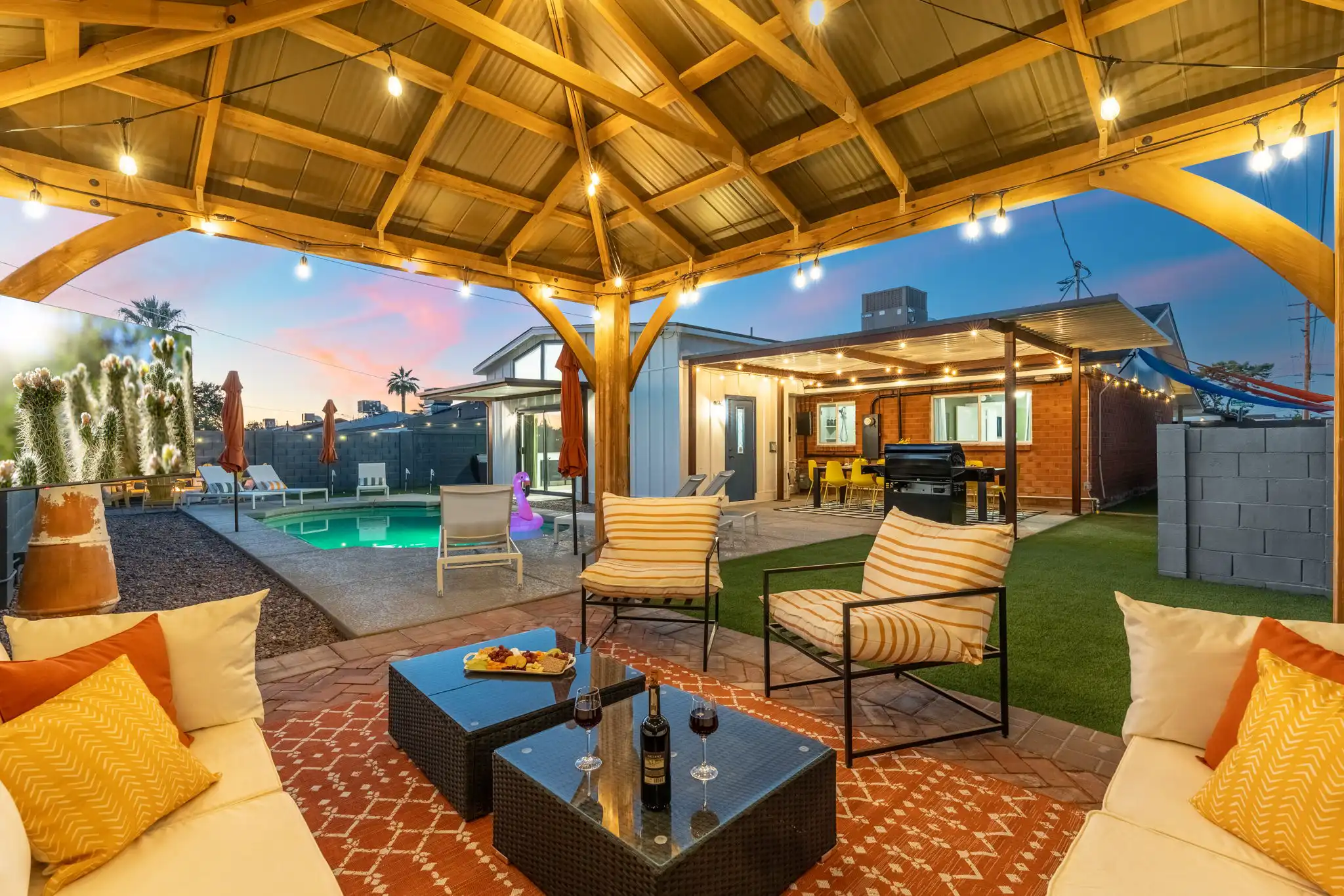 A covered wooden pergola over a brick patio with string lights, featuring a comfortable outdoor sofa, wine, and appetizers. A turquoise pool is visible in the background at sunset.