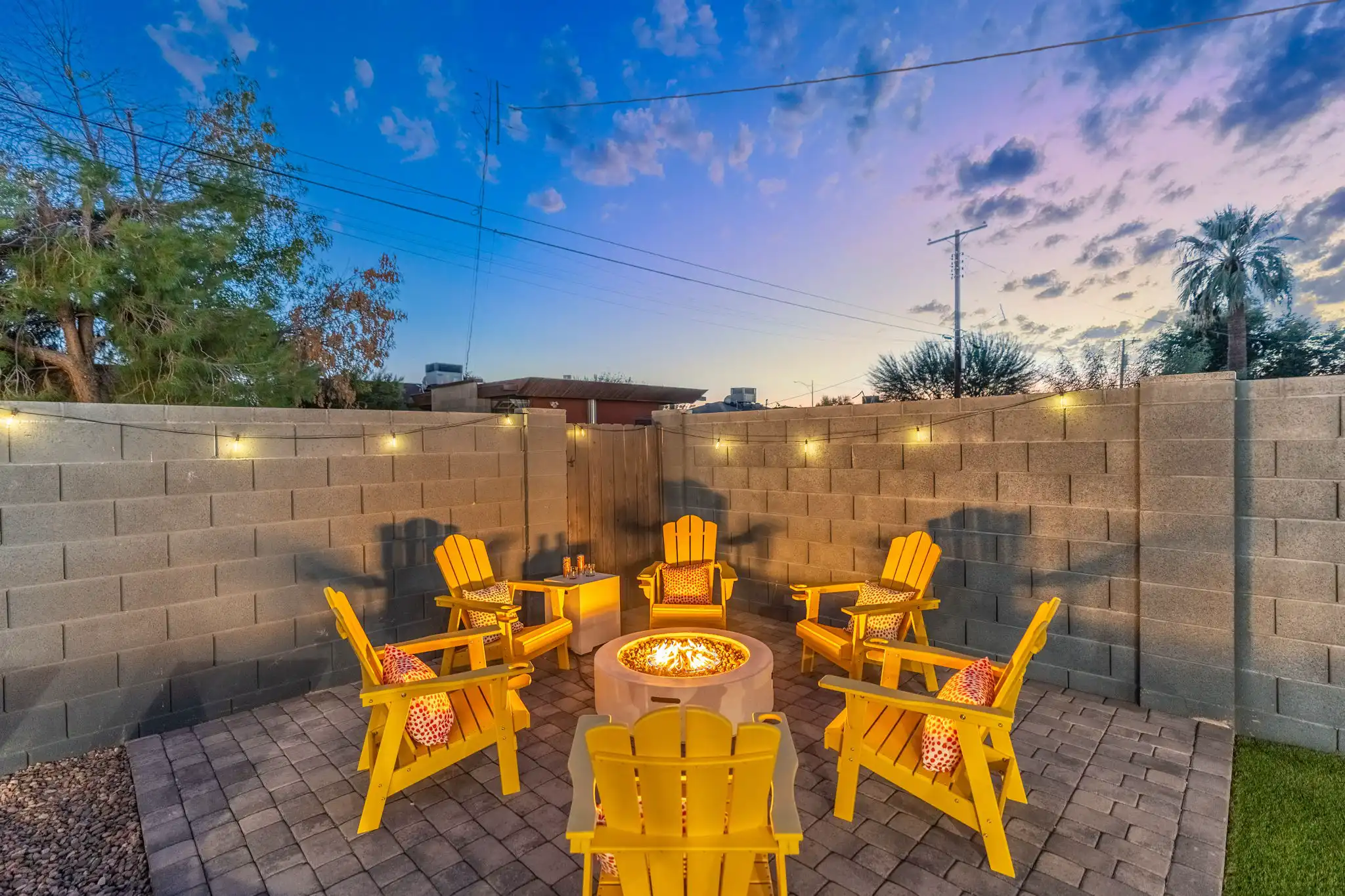 Six bright yellow Adirondack chairs arranged on a paver patio surround a lit fire pit, all set against a tall block wall at dusk.