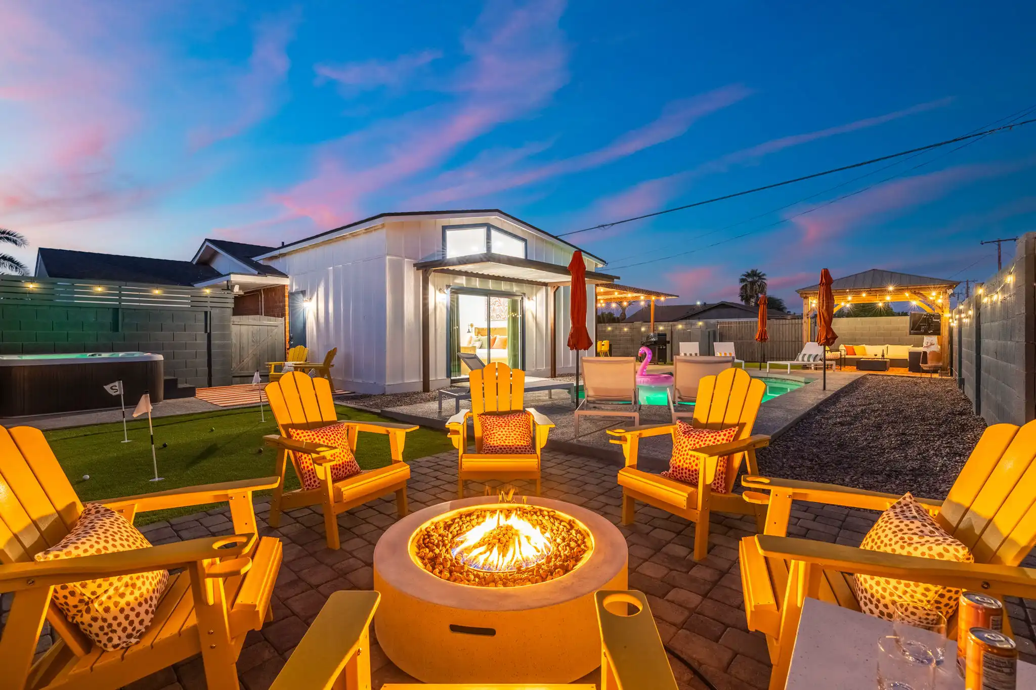 An inviting evening view of a vacation rental backyard featuring a glowing fire pit surrounded by bright yellow Adirondack chairs. A pool, hot tub, and guesthouse are visible.