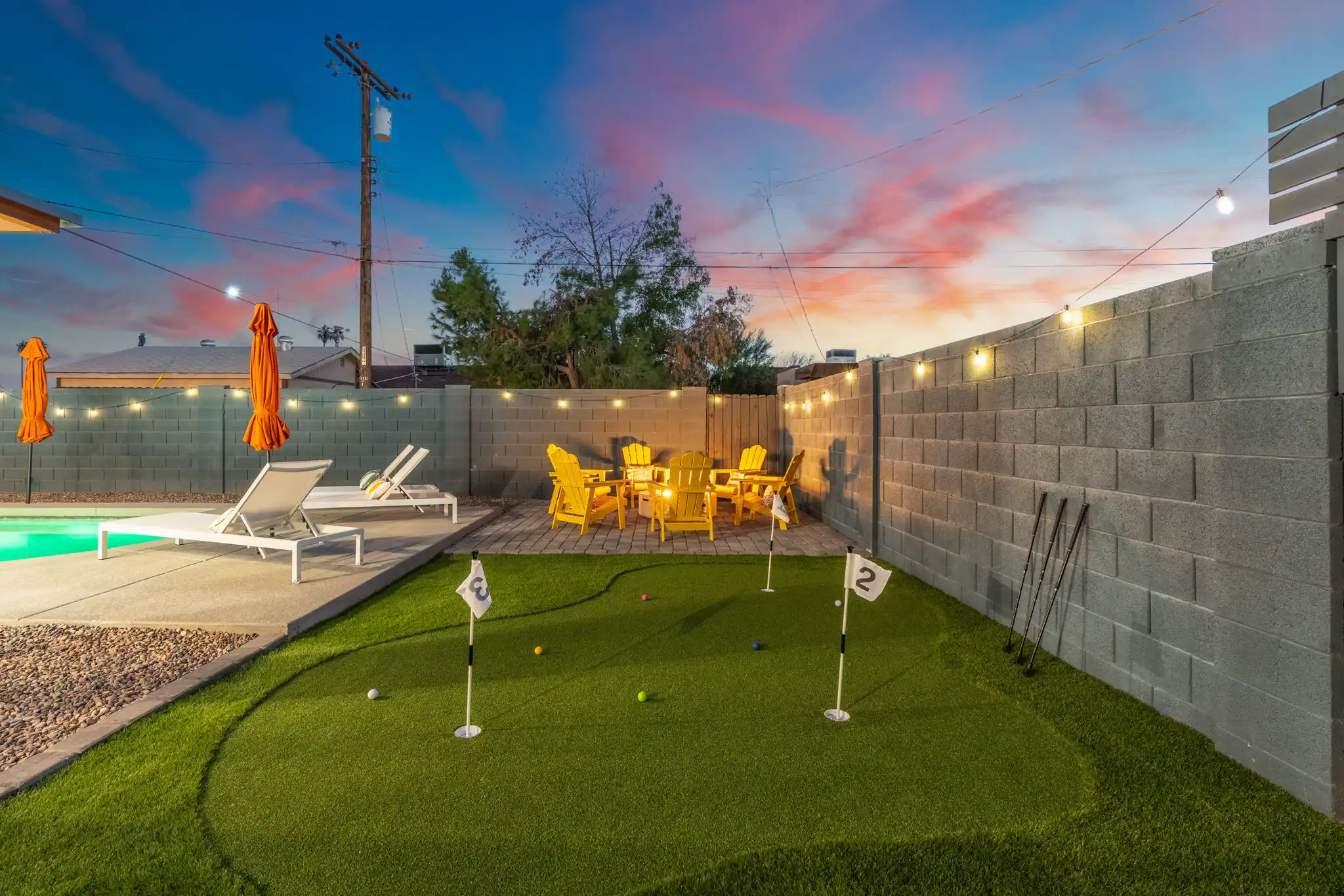 A brightly lit evening backyard featuring a mini putting green with three holes. Yellow Adirondack chairs surround a small patio area, all set against a sunset sky.