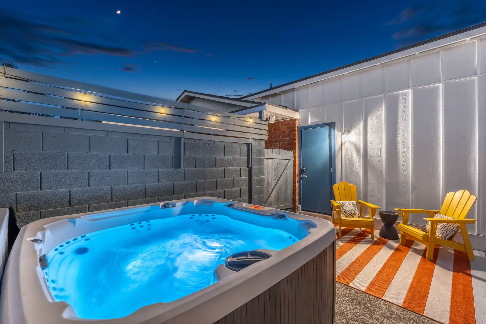 An illuminated hot tub glows bright blue on an outdoor patio at twilight, with two yellow chairs and a vibrant orange and white striped rug nearby.