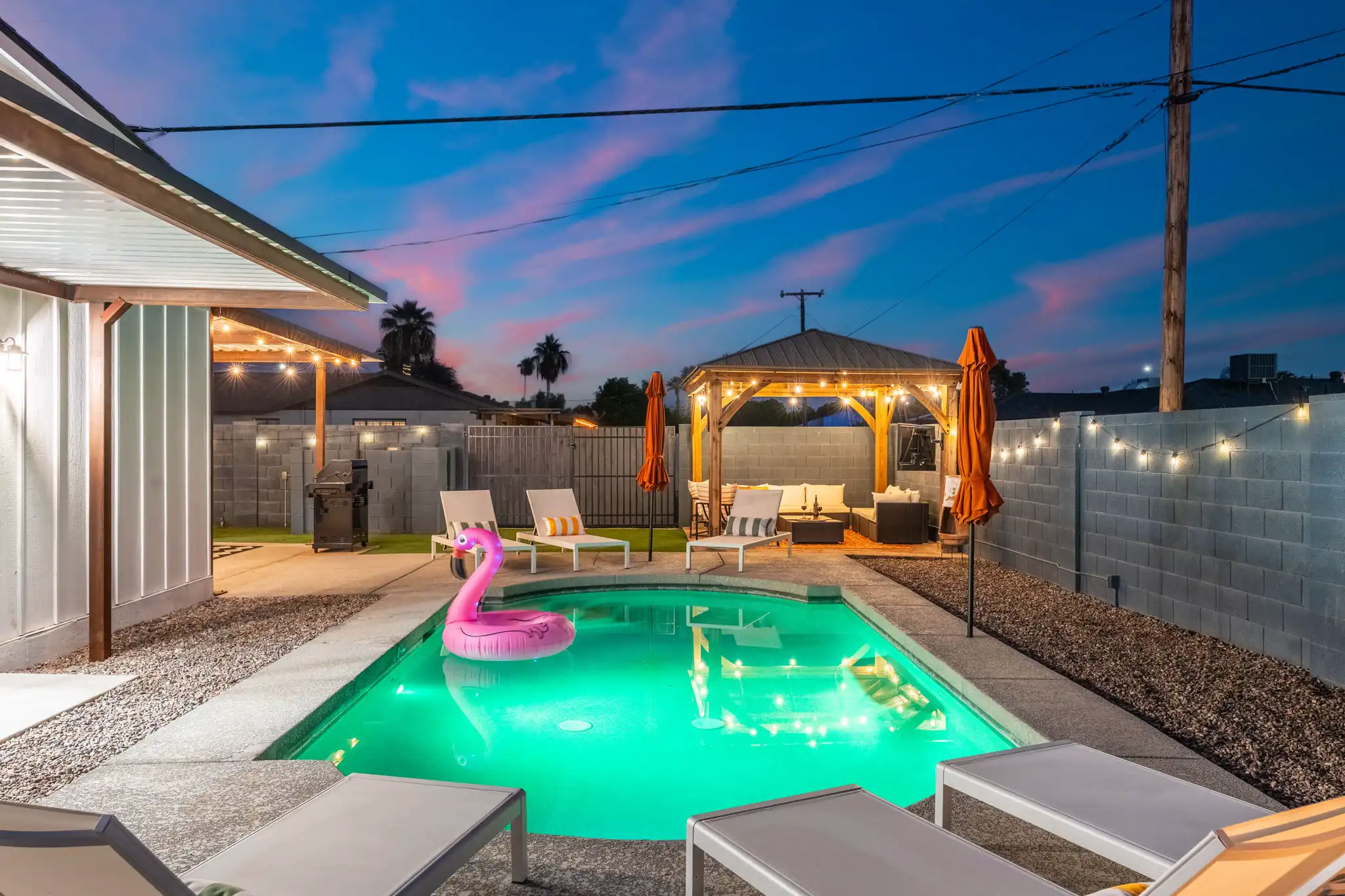 A bright green illuminated swimming pool in a backyard at sunset, featuring a pink flamingo float. Lounge chairs surround the pool, and string lights illuminate a wooden gazebo with seating.