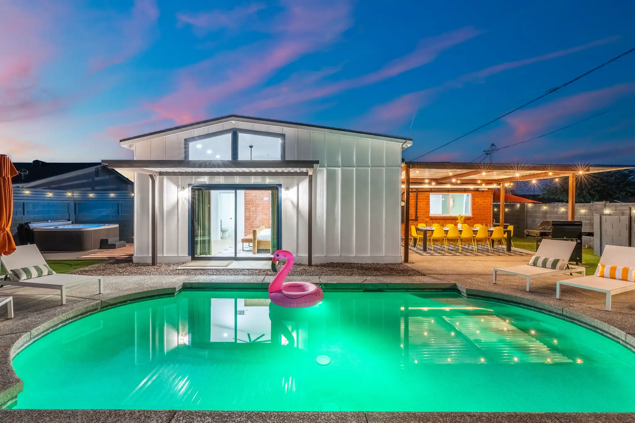 A modern vacation rental exterior at sunset, showcasing a glowing teal pool with a pink flamingo float and lounge chairs. String lights illuminate the covered outdoor dining area.
