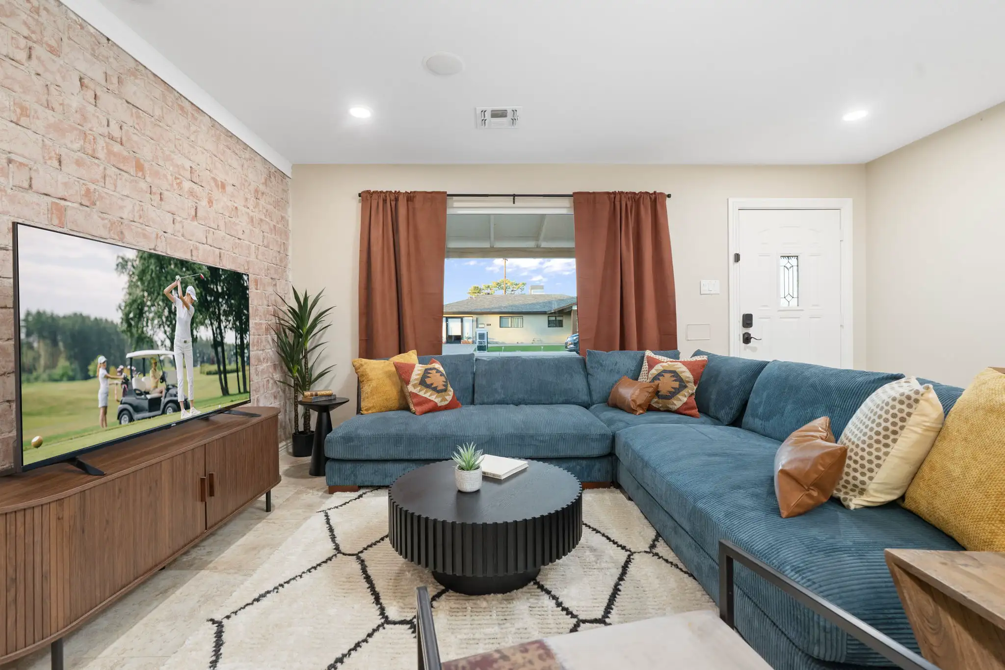 A modern, comfortable living room with a large blue corduroy sectional, a round black coffee table, and a TV next to a tan brick accent wall.
