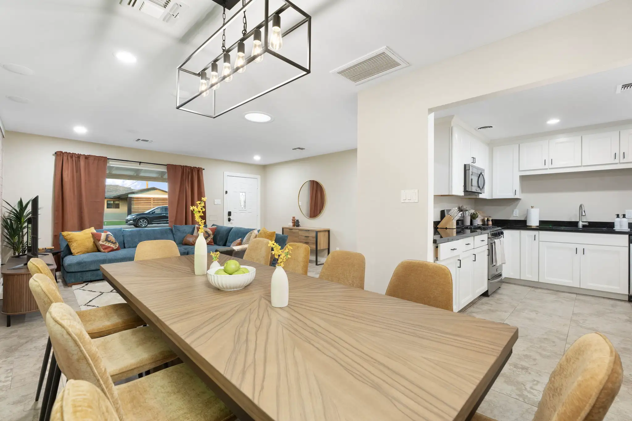 A bright, open-concept vacation rental interior showing a long wood grain dining table with mustard yellow chairs and a modern white kitchen.
