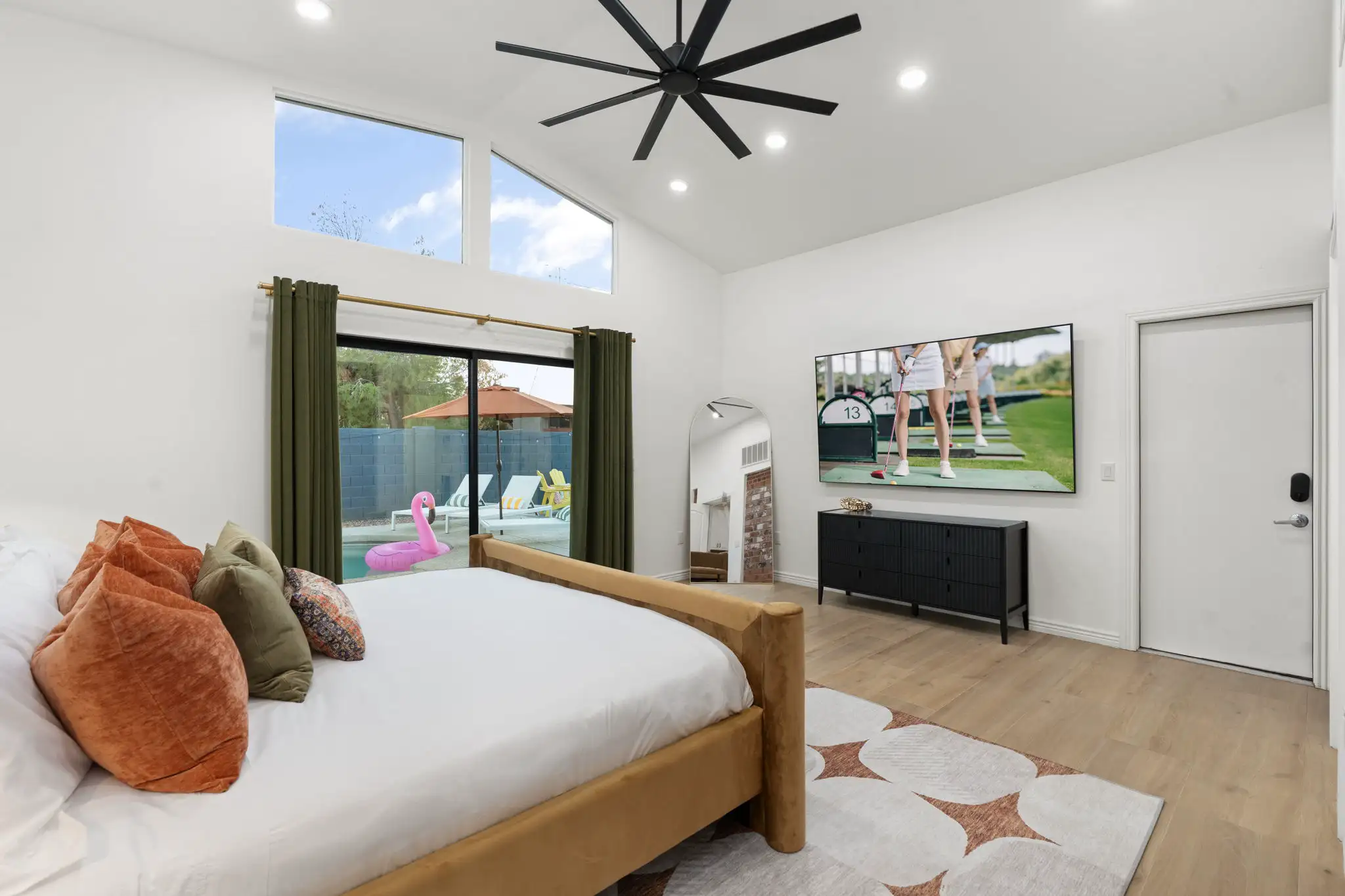 A bright, modern bedroom featuring a gold velvet bed, wood floors, and a vaulted ceiling. Sliding doors open to a pool area with a pink flamingo float.