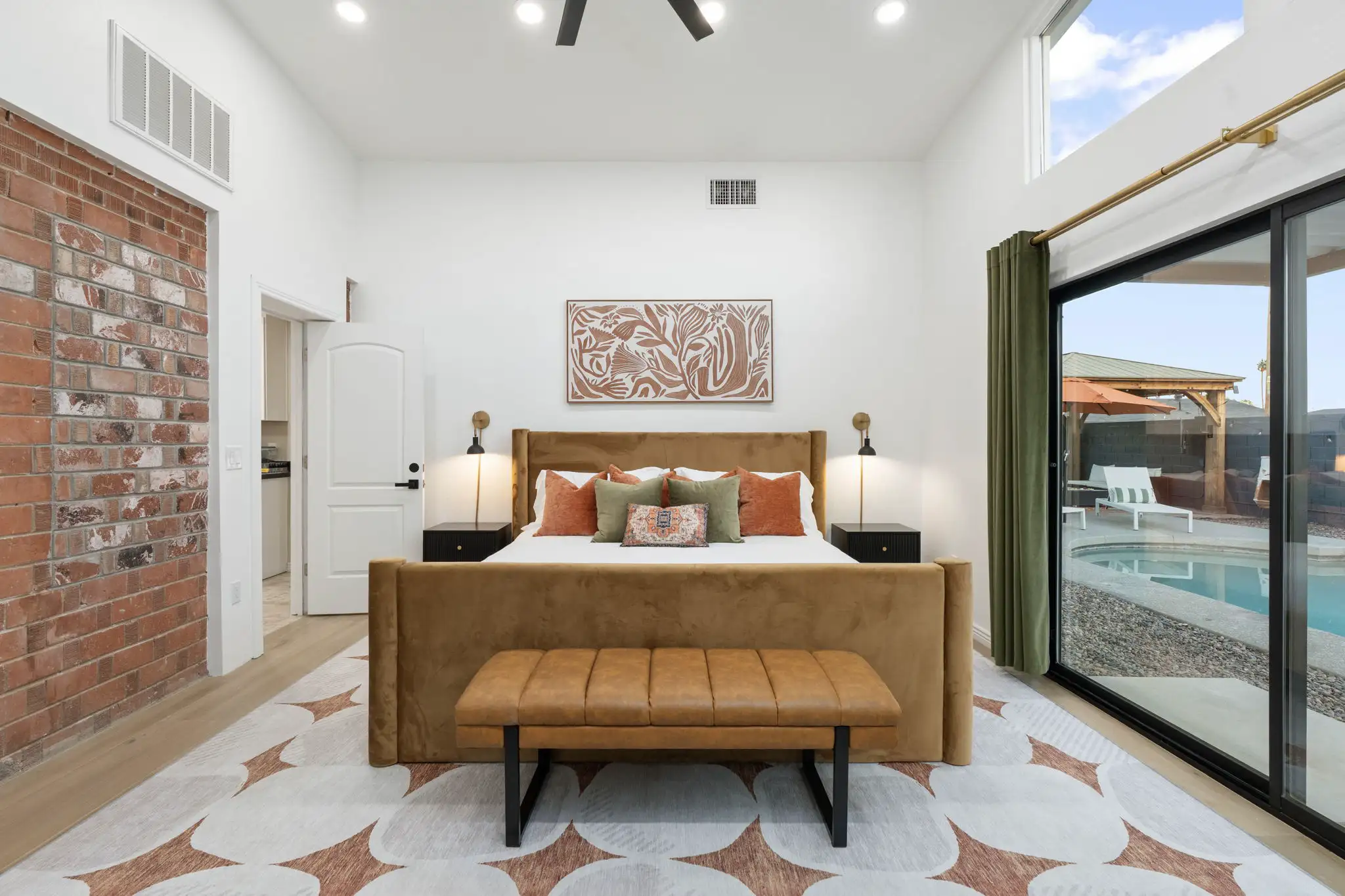 A bright, modern bedroom with a camel velvet bed and leather bench. The room has an exposed brick wall and sliding glass doors leading to a backyard swimming pool.