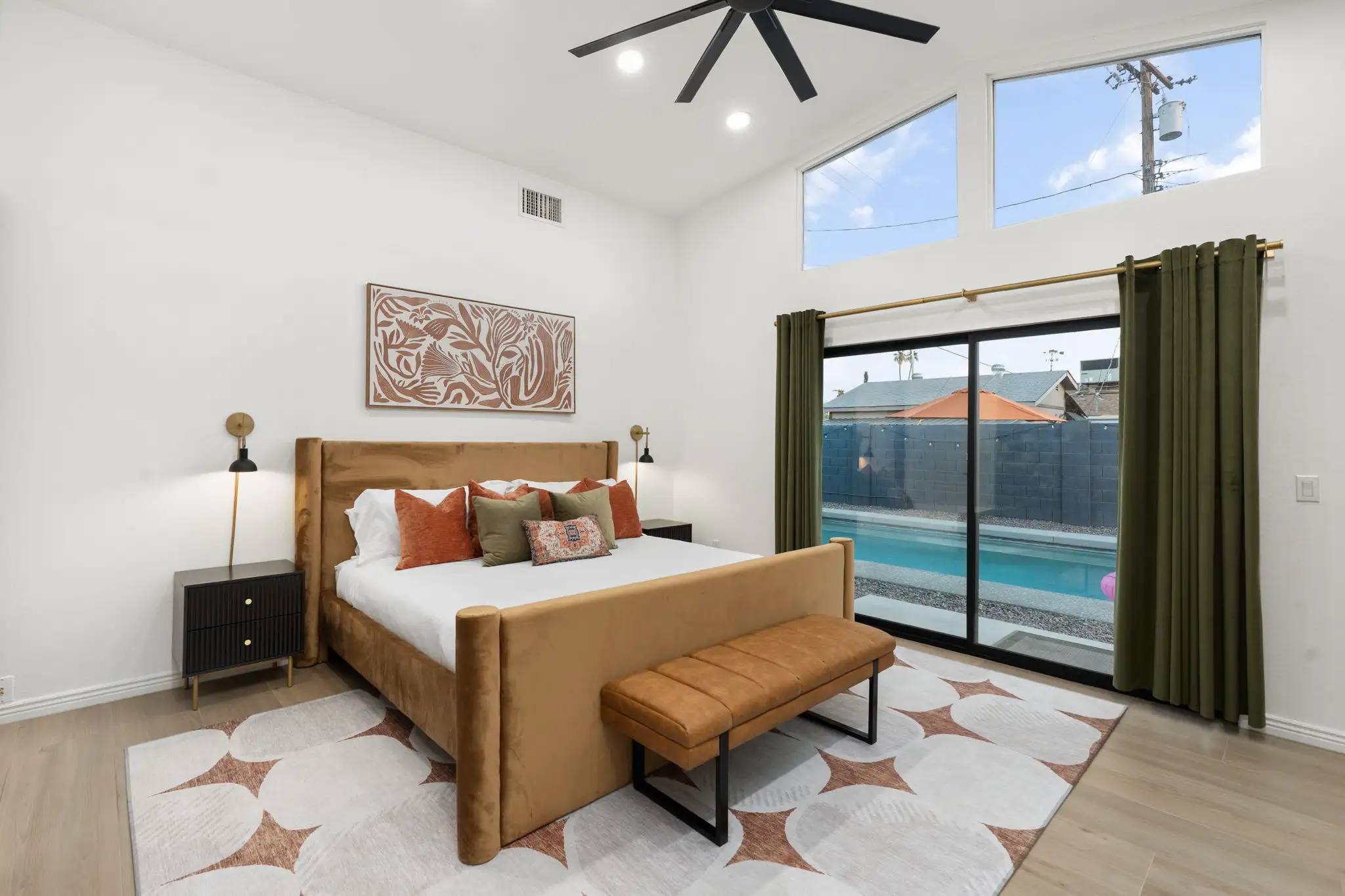 A modern bedroom featuring a gold upholstered bed, abstract art, and dark furniture. Sliding glass doors open to reveal a swimming pool and outdoor area.