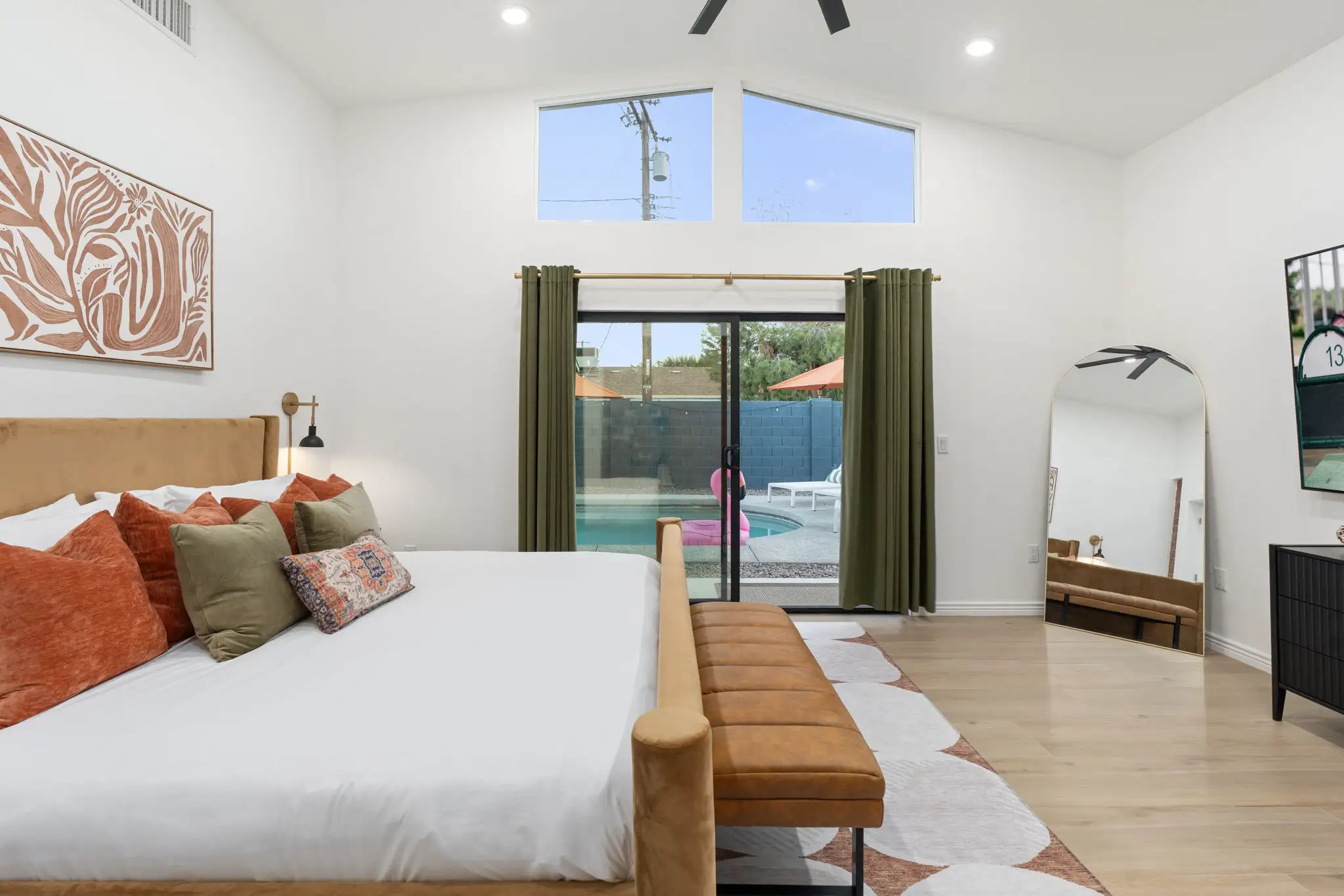 A sunlit bedroom with a tan velvet bed, abstract wall art, and a sliding glass door opening to a backyard pool area.