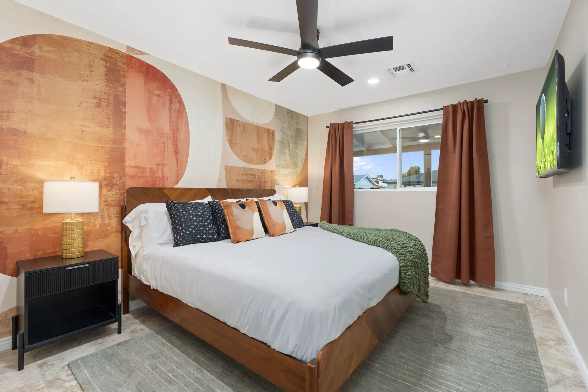 A stylish bedroom features a wooden bed set against a vibrant abstract mural wall in warm orange and tan hues, accented by a green throw blanket.